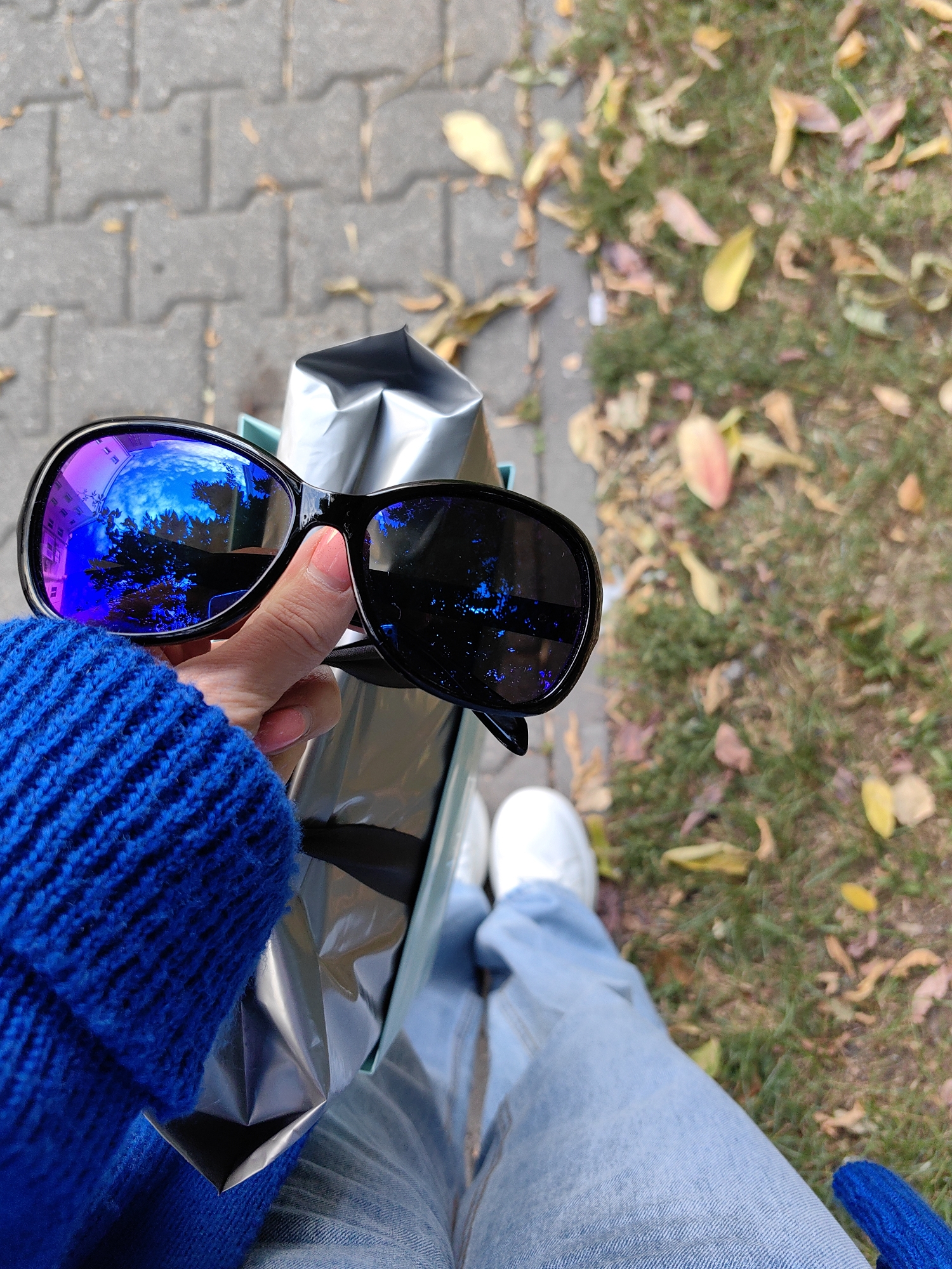 A first-person perspective shot captures a hand holding black-framed sunglasses over a shiny silver shopping bag. The sunglasses lenses reflect a bright blue sky with scattered clouds and tree branches. The background shows a paved walkway, a grassy area with fallen autumn leaves, and the lower legs and feet of a person wearing light blue jeans and white sneakers. The overall scene suggests a casual outdoor setting.
