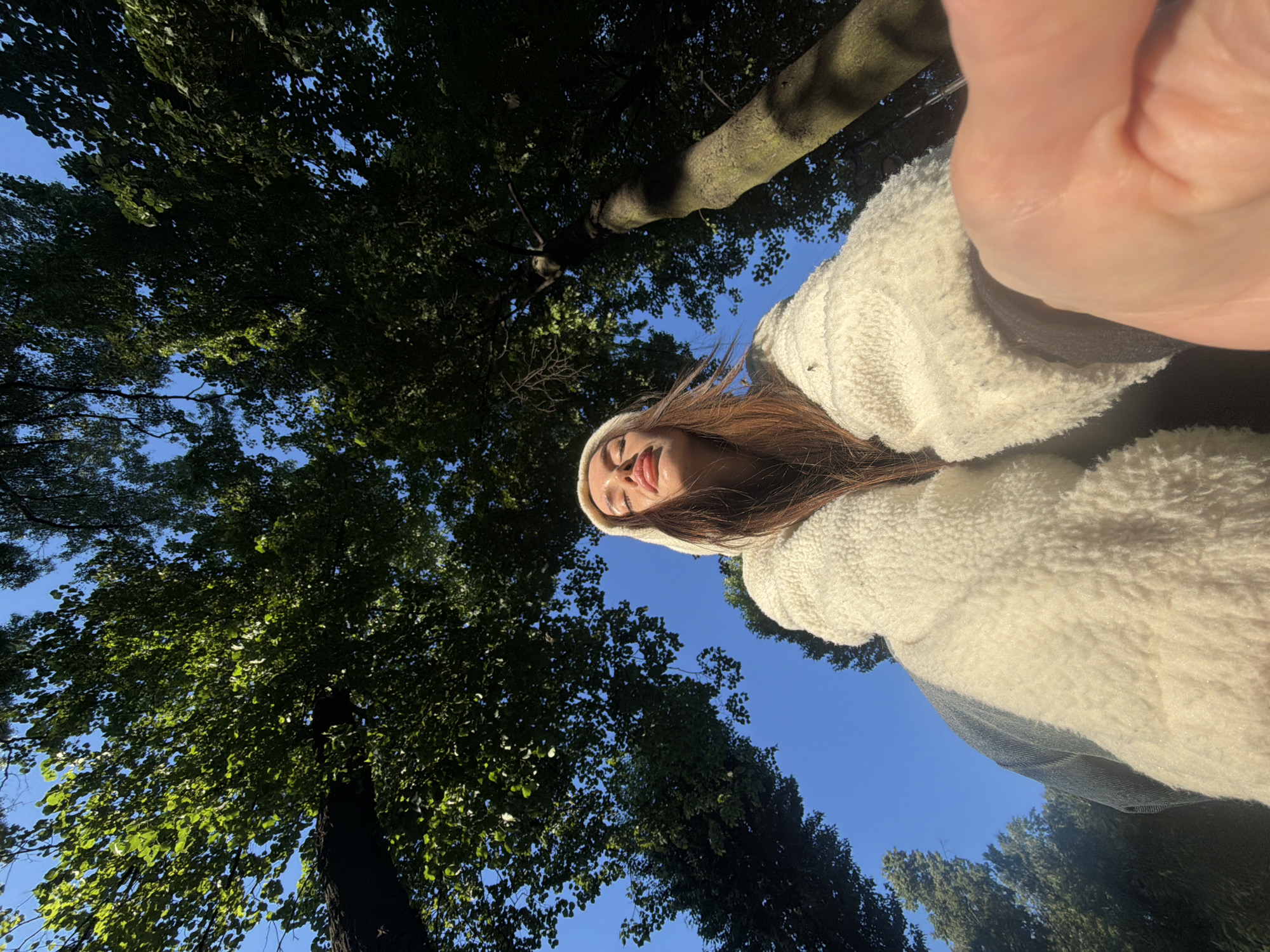 An extreme low-angle, wide shot captures a young woman posing for a selfie amidst tall trees and a clear blue sky. The woman is positioned in the foreground, facing upwards towards the camera, with her eyes closed and her lips slightly parted. She is wearing a fluffy white hooded jacket and a white beanie. Sunlight illuminates her face from above, creating a warm glow. The background is dominated by the lush green foliage of trees that stretch upwards and outwards, their branches obscuring parts of the sky. The image appears to be taken outdoors, likely in a park or forest setting. There are no discernible city landmarks visible.