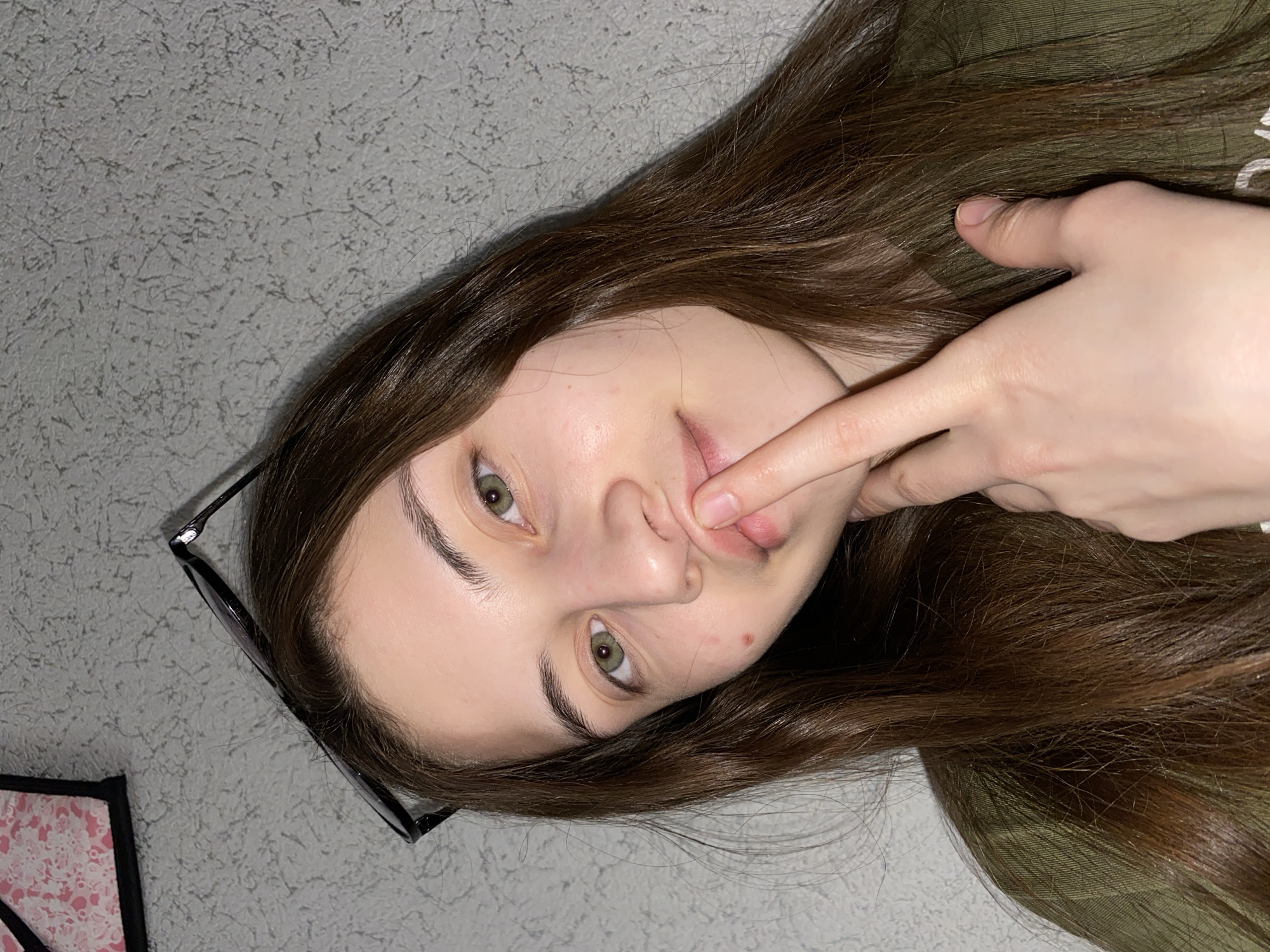 An angled, close-up indoor portrait features a young woman with long, straight, dark brown hair and light green eyes, looking directly at the viewer. Her head is tilted significantly to the left. Her right hand is raised, with her index finger lightly touching her upper lip, just above the philtrum, and her thumb resting near her jawline. A pair of black-framed glasses are visible near her left temple, resting on her hair. She has fair skin, with a few minor blemishes visible on her chin. Her expression is neutral and direct.
She is wearing what appears to be a dark green top, partially visible on the right side of the frame. The background is a light gray, textured wall. In the bottom left corner, a small section of an object with a pink and white patterned surface, possibly a book or a small bag, is visible. The lighting suggests an indoor setting, likely artificial. There are no visible location cues within the image itself to confirm the provided context of Votkinsk, Russia. No text is visible in the image.