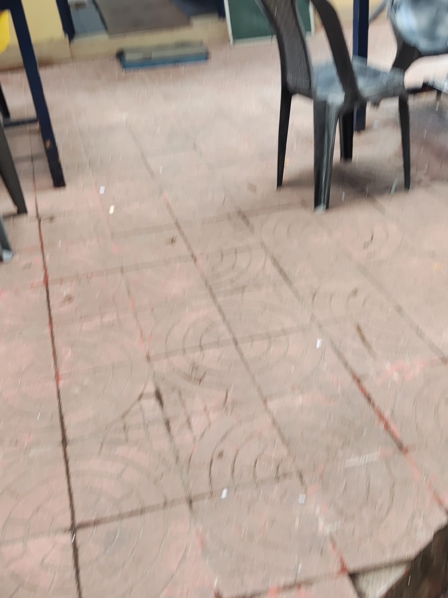 The image depicts a section of tiled flooring, likely indoors, with two chairs visible. One chair is black plastic, and the other chair is blue. The location is specified as Haini, India, although no specific city elements are visible to confirm this. There are no people visible in the frame. The overall scene is an interior setting featuring basic furniture and flooring.