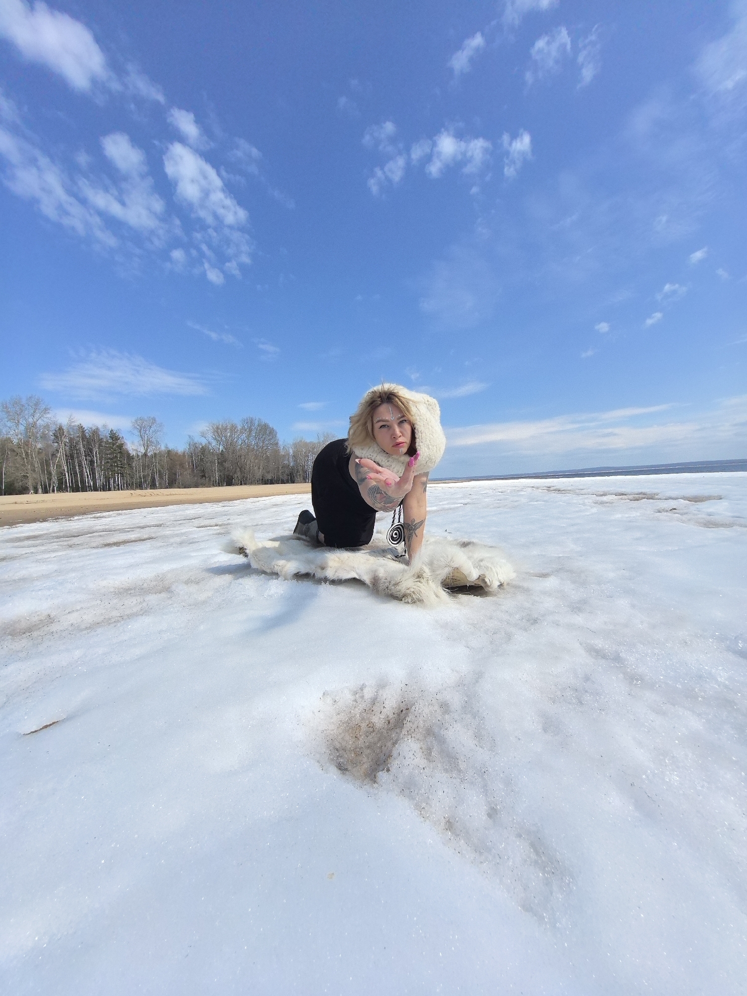 A woman poses on a vast, snow-covered landscape under a bright blue sky with scattered white clouds. She has blonde hair and is wearing a white furry hood or scarf, a black top, and black pants. She is kneeling on a white furry rug or pelt, leaning forward, and extending her right hand towards the camera in a gesture resembling a blown kiss. Tattoos are visible on her arms and shoulders, and she has white markings on her forehead.
The setting appears to be a large, partially frozen body of water, possibly a lake or extensive river, given the wide horizon. The foreground is covered in melting snow and ice, with patches of brown earth or dirt showing through. In the background, a line of bare deciduous trees stands on a sandy or dirt bank, bordering the water. The weather is sunny and clear, suggesting a cold, bright day, likely in late winter or early spring in a location like Noviy, Russia. No visible text is present in the image.