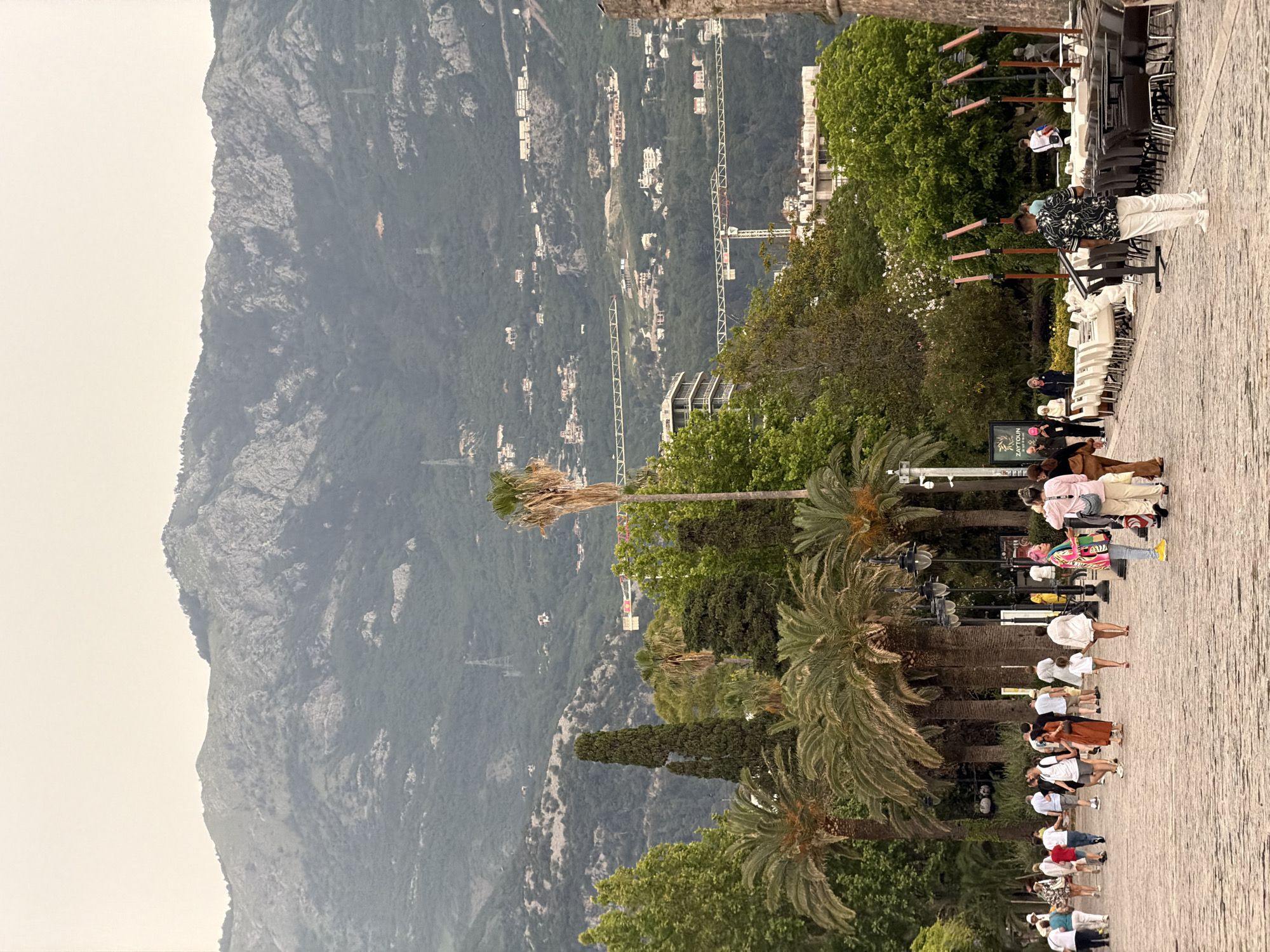 The image shows a street scene in Budva, Montenegro. A large mountain dominates the left side of the frame, with buildings and construction cranes visible on its slopes. The right side of the image features a paved walkway lined with palm trees and other greenery. People are walking along the walkway, and there are tables and chairs set up, suggesting a restaurant or cafe area. A man in a patterned shirt stands near the tables.