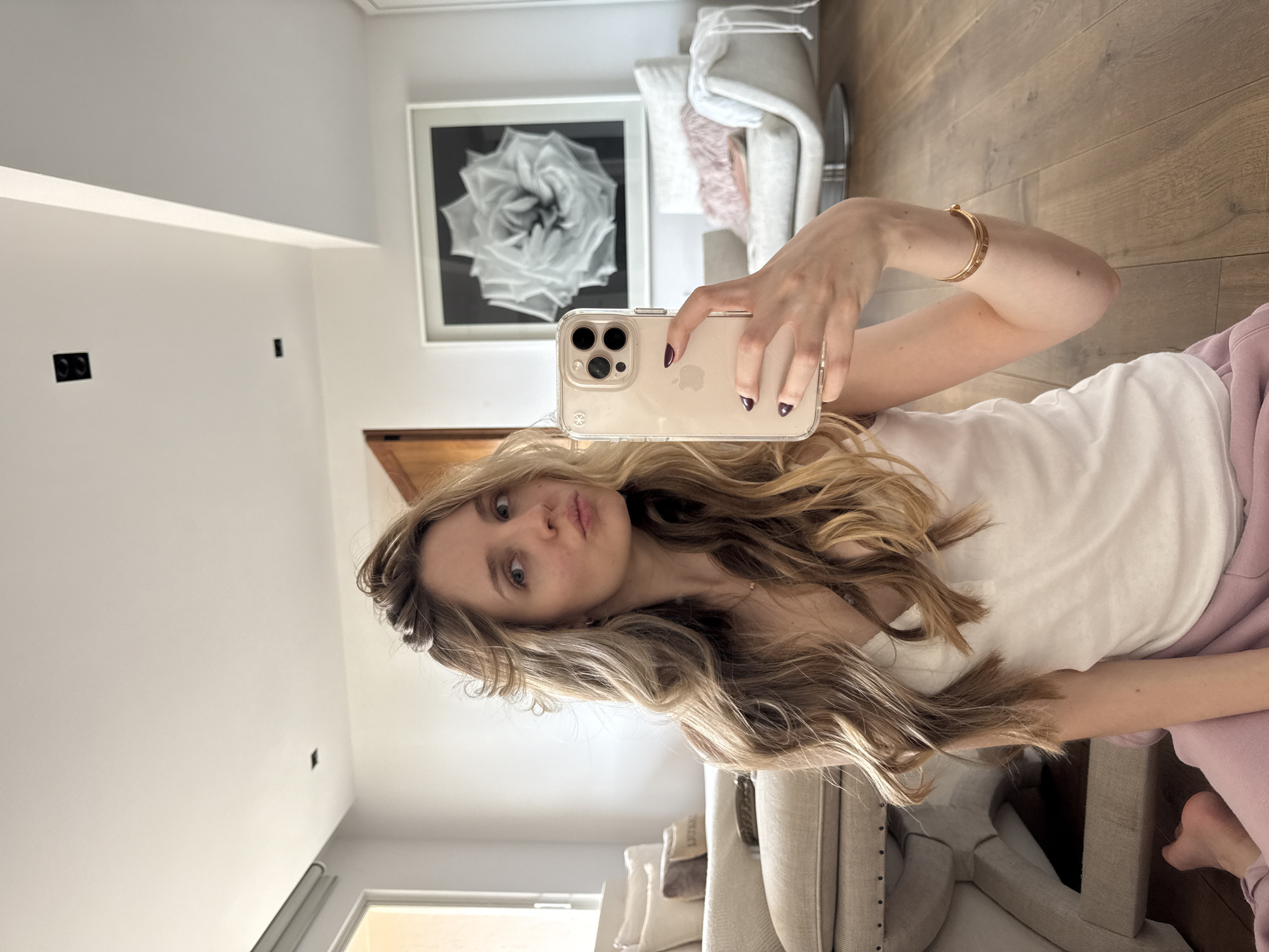 The image is a mirror selfie of a young woman taken indoors within a brightly lit, modern setting. The woman has long, wavy blonde hair and blue eyes, and is captured with a slight pouting expression. She is dressed casually in a white t-shirt and light pink sweatpants. Her left hand, adorned with dark nail polish, holds a light-colored iPhone in a clear case, showcasing the visible Apple logo and its triple camera system. A slender gold bracelet is worn on her right wrist.
The background reveals a contemporary interior space. White walls and warm wooden flooring are prominent. A large, square framed artwork depicting a detailed white rose against a black background hangs on one wall. Part of a white upholstered armchair, featuring a pink fluffy pillow, is visible next to the artwork. Another upholstered furniture piece is partially seen in the lower right. The room is well-lit, indicating it is likely daytime. Small black electrical outlets or fixtures are present on a white wall. The scene suggests a comfortable, stylish home environment, located in Mexico City, Mexico.