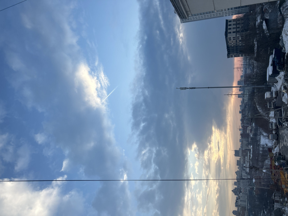 The image displays an urban panoramic view of Perm, Russia, under a partially cloudy sky. The sky exhibits a gradient from deep blue at the top to lighter hues and a warm, yellowish-orange glow near the horizon, indicative of either sunrise or sunset. A distinct white contrail from an aircraft is visible diagonally in the upper right quadrant of the sky.
A thin, dark vertical cable or wire runs along the entire left edge of the frame. In the mid-ground, a tall, slender cellular communications tower, equipped with multiple antennas, stands prominently. Below the tower and extending across the lower portion of the image, there is an industrial or storage area characterized by snow-covered ground and numerous parked vehicles, including vans and trucks. Bare trees and utility poles with power lines are interspersed throughout this area.
The background features a sprawling cityscape composed of various multi-story buildings, including a large, dark-colored structure on the right side and a tall, light-colored high-rise building adjacent to it. Snow is evident on the ground and various surfaces, suggesting cold weather conditions. No people or active interactions are discernible within the scene.