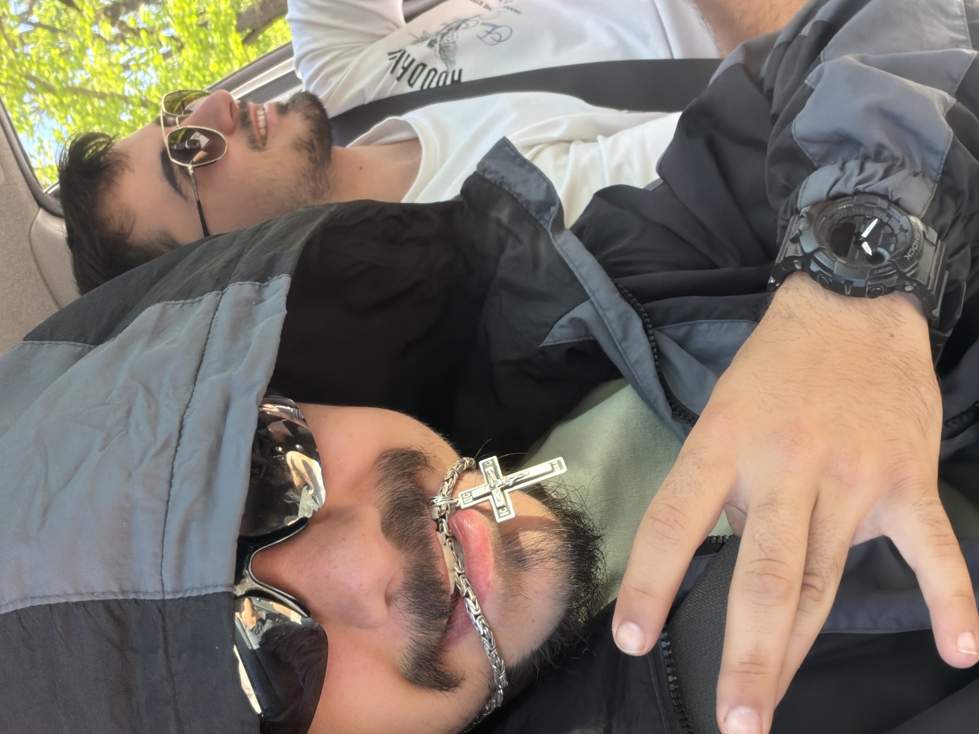Two men are sitting in a car. One man is wearing sunglasses and a silver cross necklace. He is wearing a gray and black jacket. The other man is wearing sunglasses and a white t-shirt. A hand wearing a black watch is visible. The location is Unknown, Unknown.