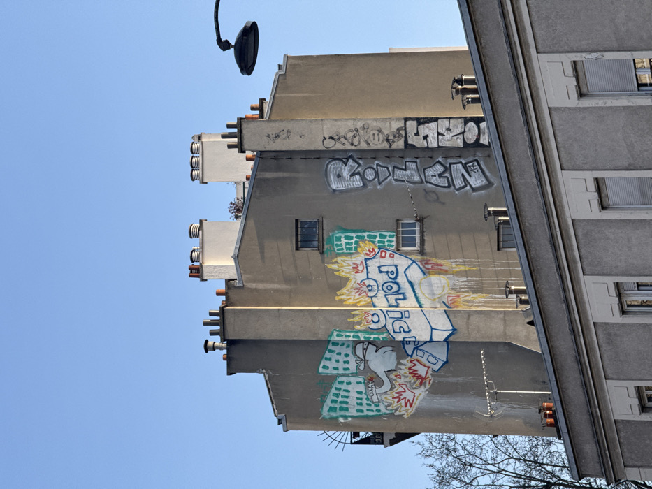 An exterior view captures a multi-story building facade in Paris, France, under a clear blue sky. The central subject is a beige-gray residential or commercial building featuring numerous dark-paned windows of varying sizes and multiple chimneys on its roofline. A prominent mural on the left section of the facade depicts a stylized, hooded figure, possibly a person, in white and gray, throwing an object resembling a Molotov cocktail at a flaming, block-lettered "POLICE" vehicle. Green outlines of urban buildings are visible behind this scene. Yellow, orange, and red flames emanate from the "POLICE" vehicle, with paint drips extending downwards. To the right of this mural, a vertical section of the facade displays large, gray and white graffiti spelling "ROLLAN," with additional illegible tags below. Further right, more black graffiti appears vertically. Above, the building's roof features several white-painted chimney stacks with ornate tops and smaller terracotta-colored chimneys. A black, curved street lamp extends into the frame from the upper right. Below the main building, the upper portion of a distinct, gray building with white-framed windows and closed roller shutters is visible. Bare tree branches are present on the lower left.