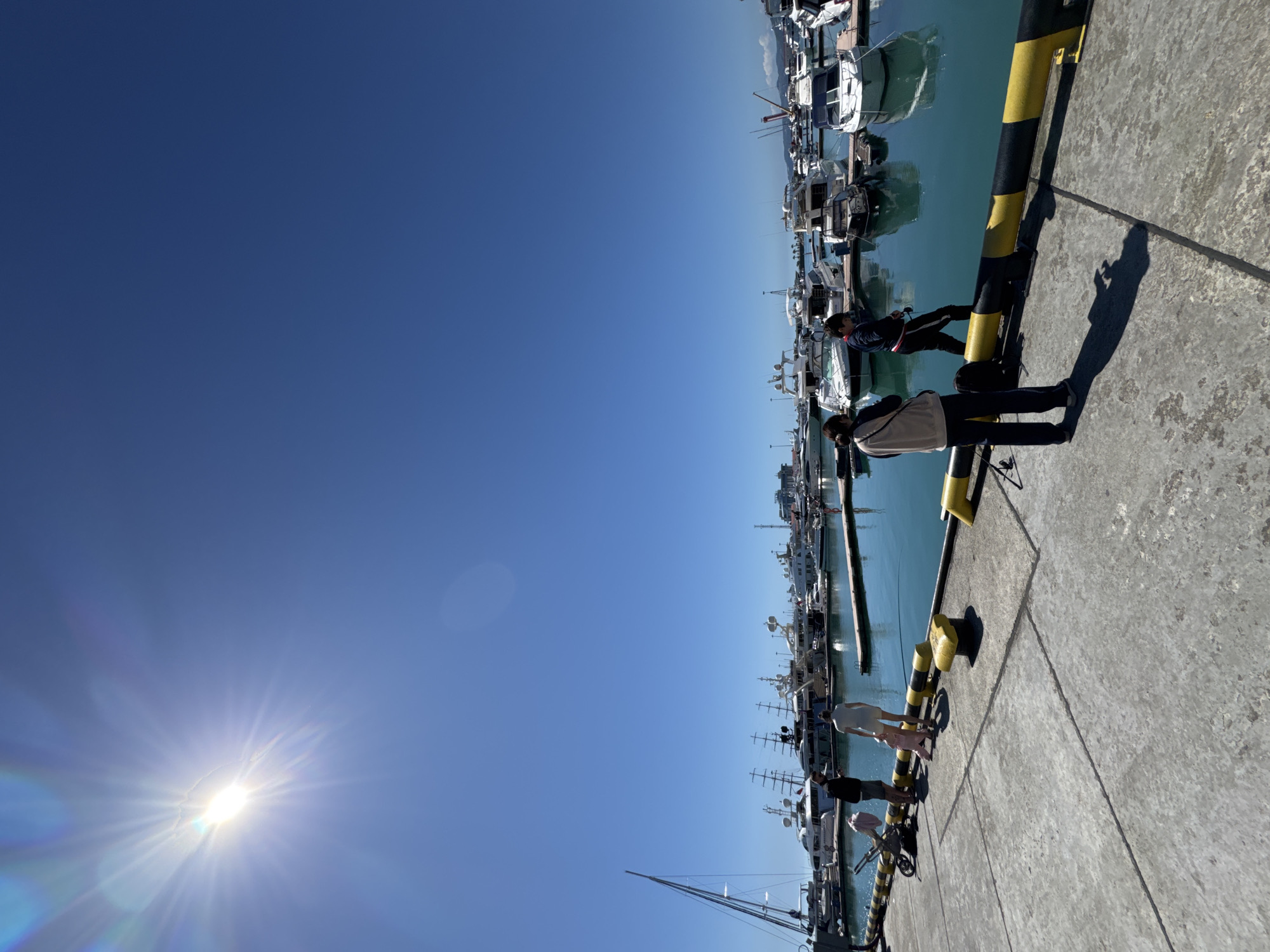 This image captures a vibrant, sunny day at a bustling marina, likely in Sochi, Russia. The sky, a brilliant expanse of clear, deep blue, dominates the upper portion of the frame. The sun, a powerful light source, is prominently visible in the upper-left corner, emitting a strong starburst and lens flare that creates colorful refractions.
In the foreground and extending along the right side of the image is a wide concrete pier or dock. This dock is clearly marked by several distinctive yellow and black striped bollards. Numerous individuals are scattered along the dock. In the mid-ground, two people stand near the bollards, appearing to observe the boats or the surroundings. One person, dressed in a light-colored jacket and dark pants, holds what looks like a camera or phone, possibly capturing the scene. Another individual, wearing a dark jacket with a reddish trim, stands nearby. Further down the dock, additional figures, including what appear to be children, are visible, some near a bicycle or scooter.
To the left of the dock, the calm, blue-green water is filled with a multitude of boats and yachts. These vessels, predominantly white and light-colored, vary in size and design, featuring cabins, various antennae, and masts, indicative of both leisure and possibly some fishing activities. The distant horizon, seen above the boats, reveals a subtle outline of land or structures against the clear sky.
The bright mid-day sun casts sharp, elongated shadows of the people and bollards onto the concrete surface of the dock, suggesting the sun is at a considerable angle, possibly late morning or early afternoon. The water surface is still, reflecting some of the boats and the sky. No visible text is discernable within the image. The scene conveys a relaxed and picturesque atmosphere of a lively harbor on a beautiful day.
