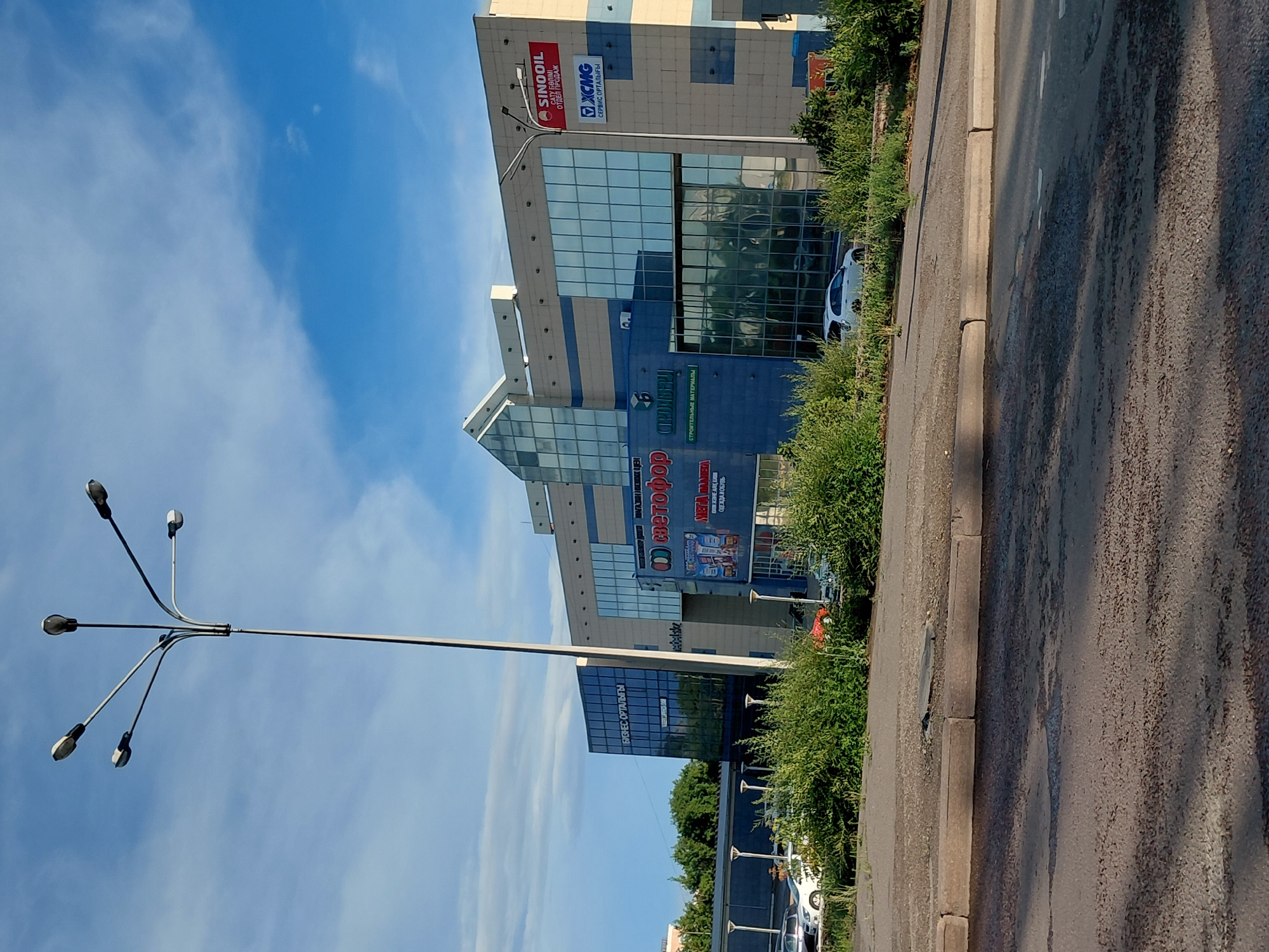 The image shows a street view of a large, modern commercial building in Qarağandı city administration, Kazakhstan. The building features a mix of blue and beige colored panels and extensive glass windows. Several signs are visible on the building, including "SINOIL", "XCMG", "МЕТАФОР", and "СТРОИТЕЛЬНЫЕ МАТЕРИАЛЫ". In front of the building, there is a parking lot with several cars, and lush green bushes and trees line the area. A tall lamppost with multiple lights stands in the foreground, against a bright blue sky with scattered clouds. The street in the foreground appears to be paved with asphalt.