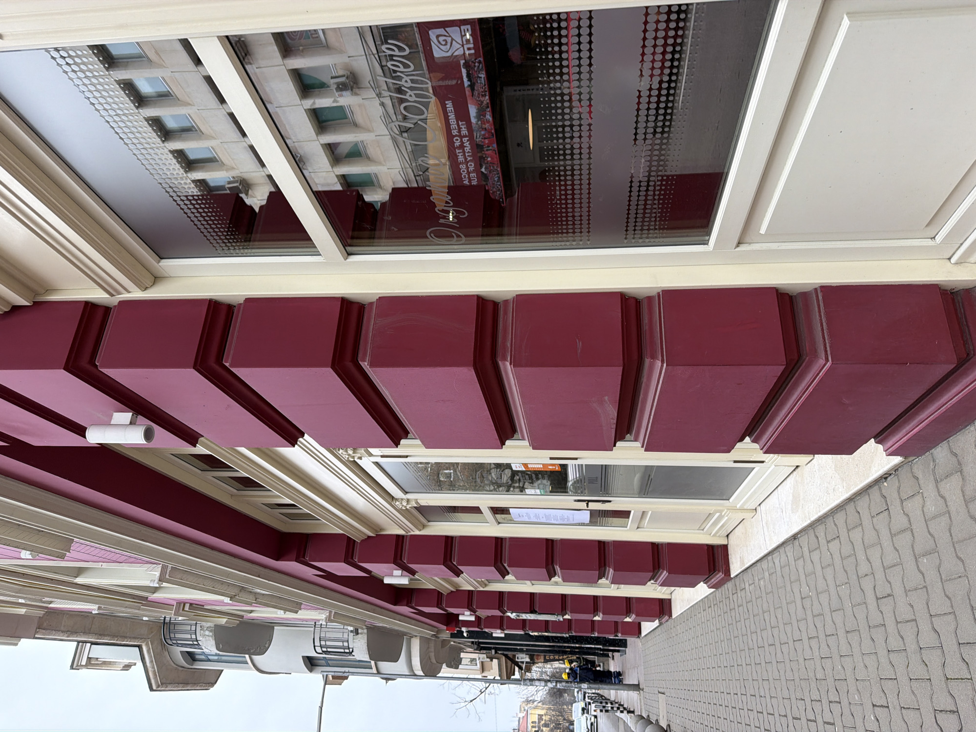 The image shows the facade of a building in Sofia, Bulgaria, viewed from below. The building has a series of dark red, projecting elements below a window. The window reflects a building across the street, and a sign that reads "Coffee Org." Workers in yellow vests are visible on the street level. The sidewalk is paved with gray bricks.
