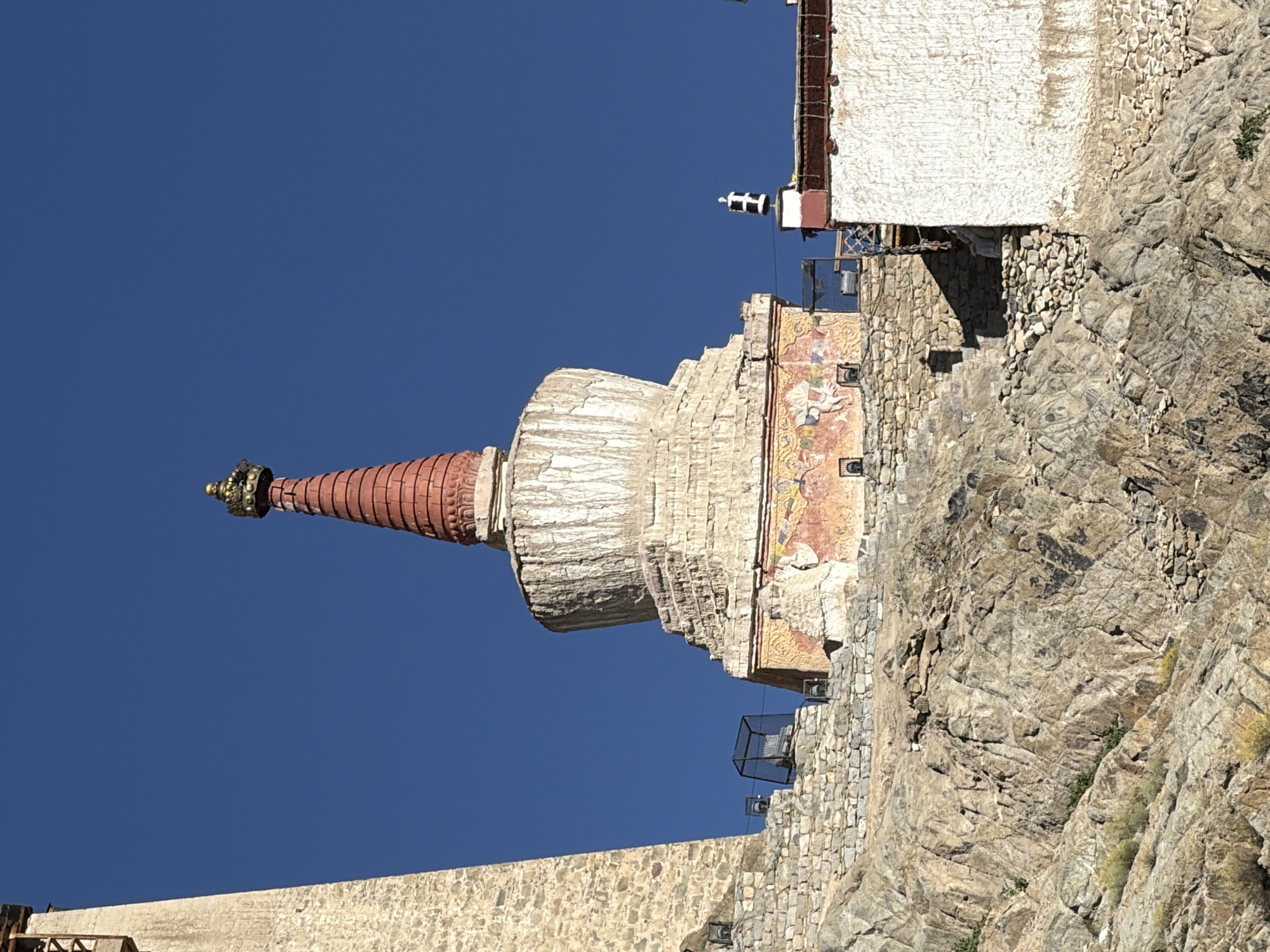The image captures a stupa and surrounding structures atop a rocky hill against a clear blue sky. The stupa is prominent, with a red, tiered conical top adorned with a golden finial. Its white dome-like base is decorated with faded murals. Adjacent to the stupa, a tall, light-colored tower with wooden balconies stands on the left. On the right, a white building with red trim and dark decorative elements is visible. The foreground consists of a steep, rocky slope with scattered vegetation. The scene appears to be within the Shanti Stupa complex in Leh, India, given the architectural style and mountainous terrain. There are no visible people or active interactions.