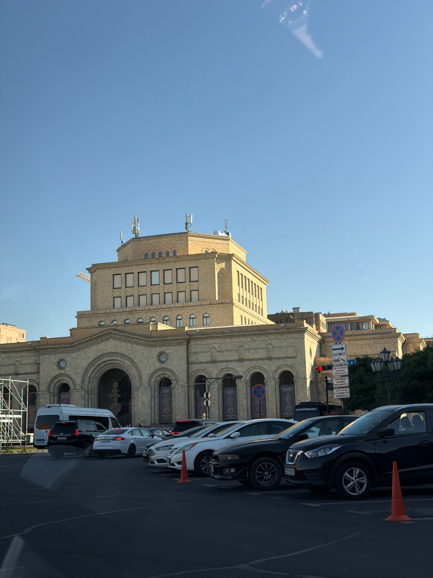 This image depicts the Republic Square in Yerevan, Armenia, under a clear blue sky. The prominent feature is the National Gallery of Armenia, a grand architectural structure made of yellowish stone. The building has a layered design with multiple windows, some appearing rectangular and others arched. Several antennas are visible on the rooftop. In front of the building, a parking lot is filled with various cars, including a white van, a black SUV, and several sedans. Some cars are parked, while others might be in the process of parking or leaving. Traffic signs are also visible on the right side of the image. The overall scene suggests a typical day in a public space with moderate activity.