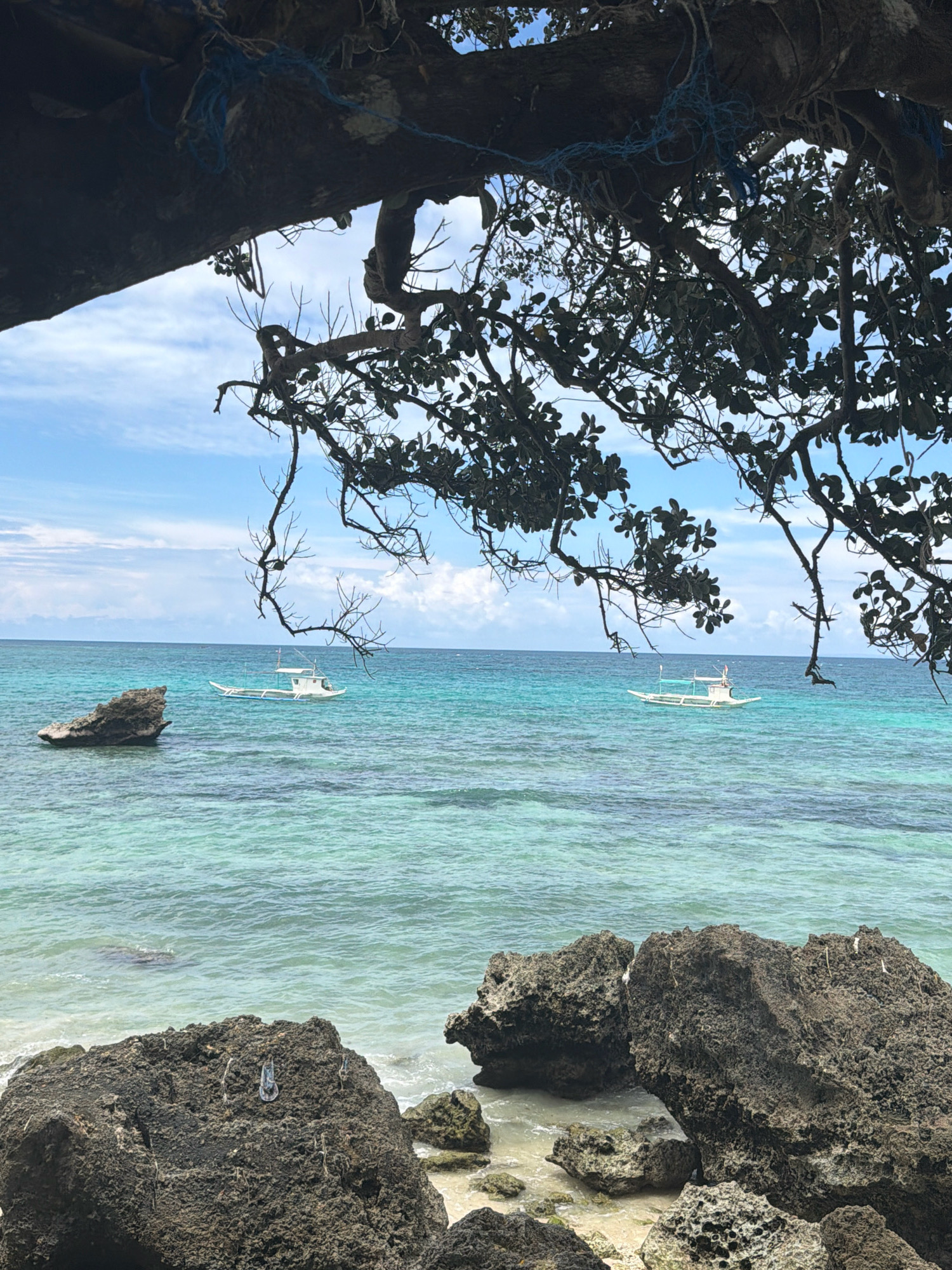The image captures a scenic view of a tropical beach in Malay, Philippines, likely Boracay Island, as suggested by the characteristic clear turquoise waters and white boats. In the foreground, dark, rough-textured rocks of varying sizes line the shore. Beyond the rocks, the crystal-clear water transitions from a light aquamarine near the shore to a deeper blue as it recedes. Two traditional outrigger boats, white with blue accents along the waterline and adorned with a canopy structure, are visible on the water. One boat is closer to a large rock formation on the left side of the frame, while the other is further out in the sea. The horizon is marked by the meeting of the sea and a bright blue sky dotted with white, fluffy clouds. A large tree branch, dark and leafy in parts and bare in others, extends from the top of the frame, partially obscuring the sky and the water, creating a natural frame for the scene. Blue rope is tied to the tree branch. The overall atmosphere is serene and idyllic, typical of a tropical island paradise.