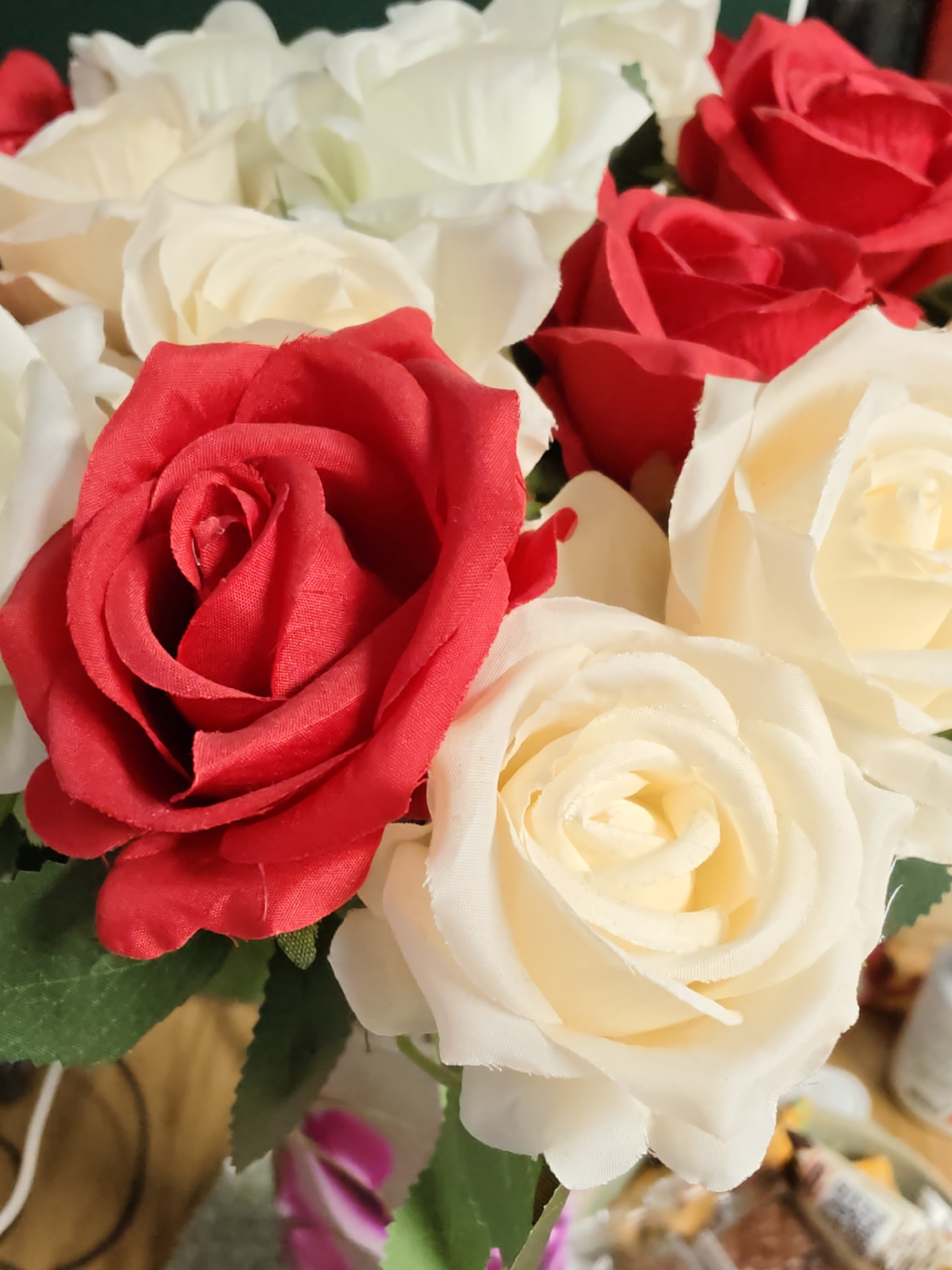 A close-up, slightly angled shot shows a bouquet of artificial roses, predominantly red and cream-colored. The roses are densely packed, with some green artificial leaves interspersed. The focus is on the flowers themselves, with their textured petals and layered arrangement. In the background, elements suggest an indoor setting, possibly a table or shelf, with hints of other objects, including what appear to be some wrapped food items or candies. The lighting is soft, highlighting the details of the rose petals. There is no information in the image to suggest a location in Petah Tikva, Israel.