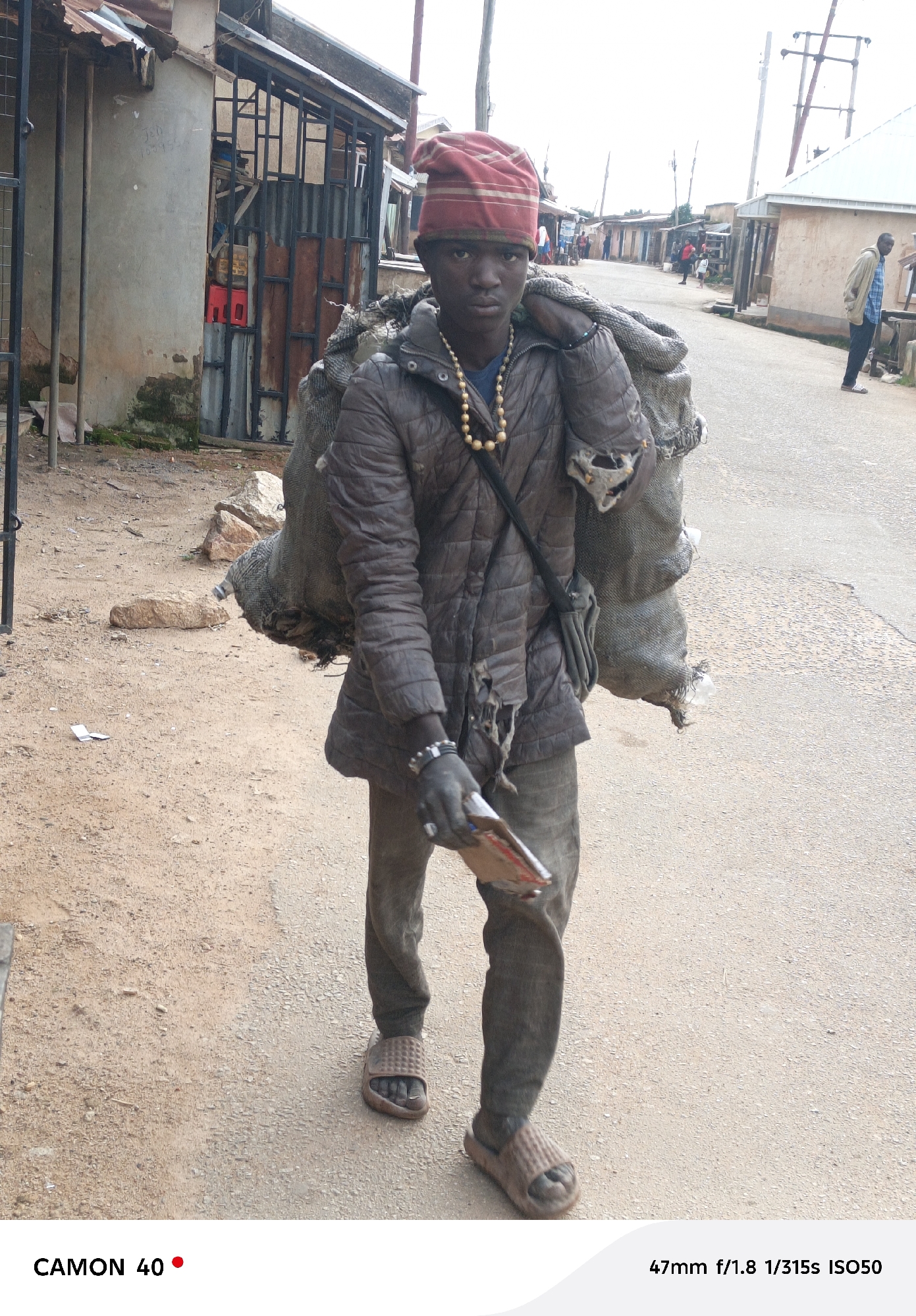 In Jos, Nigeria, a young Black male wearing a red beanie, a brown quilted jacket, dark pants, and sandals, walks down a dirt and asphalt road. He carries two large sacks of what appear to be charcoal or waste slung over his left shoulder, secured by a dark strap. The sacks are tattered and show signs of wear. Around his neck, he wears a string of large, golden beads. His right hand is clenched around a small, rectangular object, possibly a book or a stack of papers. The background shows a street lined with low-rise buildings, some with corrugated metal roofs, and utility poles. The ground is a mix of sandy dirt and worn asphalt. The scene captures a moment of daily life and labor in the urban environment of Jos.