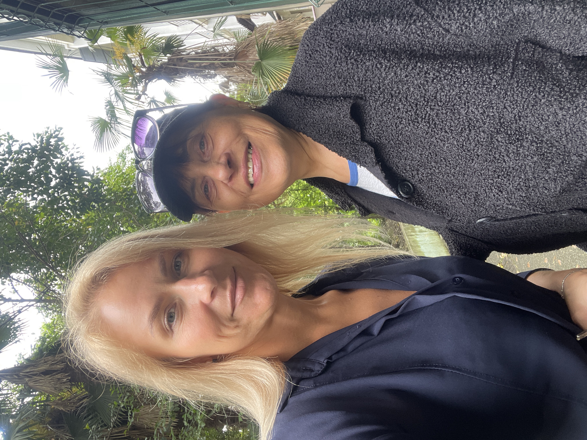 Two women are posing close together for a selfie-style photograph outdoors. The woman in the foreground has blonde hair and is wearing a dark, buttoned blouse. The woman in the background has short dark hair and is wearing a dark textured coat with a light-colored top underneath. Sunglasses are perched on top of her head. The background consists of green foliage, including palm trees, and a glimpse of a building structure. The image is taken in the city of Sochi, Russia.