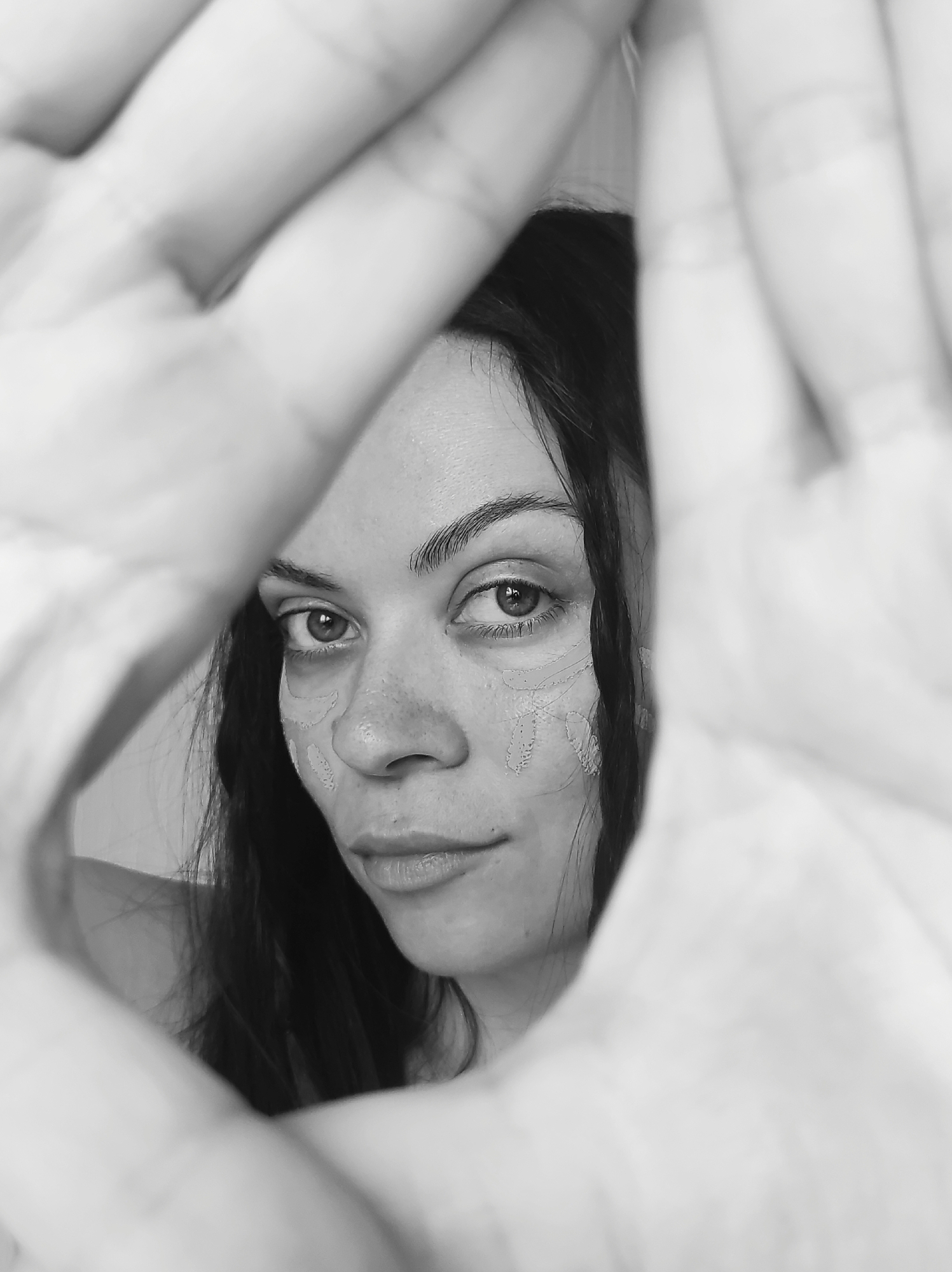 A close-up, black and white portrait features a young woman with dark hair, framed by blurred hands in the foreground. Her hands form an opening through which her face is visible, creating a sense of intimate focus. The woman looks directly at the viewer with a steady, contemplative gaze. Her dark eyes are prominent, and her eyebrows are well-defined.
Notably, there are light-colored, abstract markings or patterns on her cheeks, just below her eyes, possibly painted, drawn, or remnants of a dried cosmetic product. These marks add an artistic or unconventional element to her appearance. Her lips are slightly parted. The foreground hands are intentionally out of focus, emphasizing the sharp details of her face. The background is softly blurred and indistinct, suggesting an indoor setting.
The overall mood is artistic and introspective. The use of black and white enhances the texture and contours of her face and the surrounding hands. No specific time of day or weather is discernible from the image, and there is no visible text. The location context is Chaikovsky, Russia.