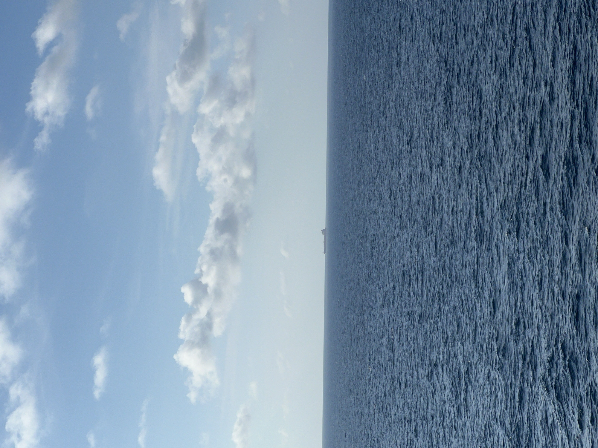 A cargo ship is visible on the horizon in the ocean under a blue sky with scattered clouds. The ocean is a deep blue with small waves. The ship is dark and appears to be a freighter. The sky is light blue and mostly clear, with some fluffy white clouds. The overall scene is one of vastness and open water.