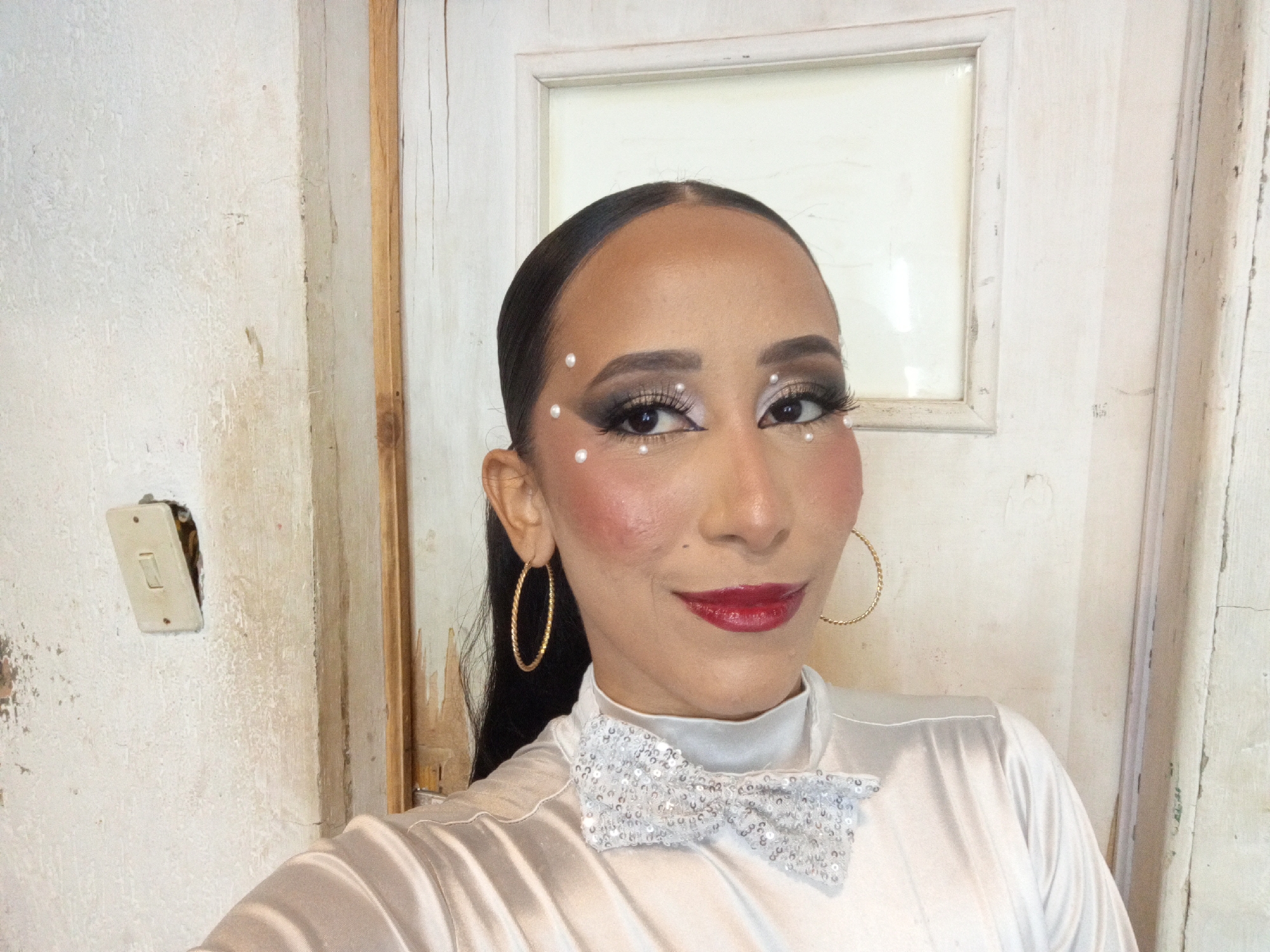 The image is a close-up selfie of a woman indoors. She has dark hair slicked back, is wearing gold hoop earrings, a white satin shirt, and a sequined bow tie. Her makeup includes dark eyeshadow, false eyelashes, red lipstick, and pearl-like decorations around her eyes.
The background consists of a textured, off-white wall with an exposed electrical switch plate and a white door with a small window inset.
The location in the city of Porlamar, Venezuela is unknown. There is no data on the photograph to indicate this location.