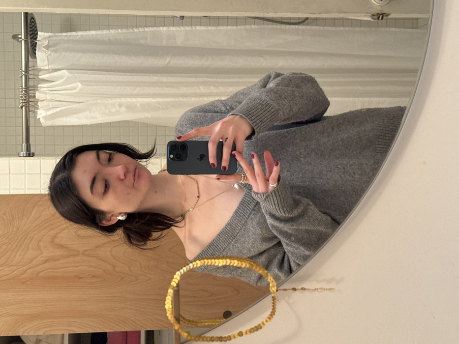 A young adult female is depicted taking a mirror selfie in a bathroom. The specific location within New York City, United States of America, cannot be determined from the provided image.
The subject has dark hair, styled loosely, and is wearing a grey V-neck long-sleeve sweater. She is adorned with silver-colored hoop earrings, multiple necklaces of varying lengths (including one with small beads and another with a larger white, irregular pendant), and several rings on her fingers, one notably a double-banded ring on her left ring finger. Her fingernails are manicured with dark red polish. She holds a dark-colored smartphone (consistent with an iPhone model) in her right hand, aimed towards the mirror. Her facial expression appears slightly contemplative or pouting, with her gaze directed towards the phone.
The background, reflected in the mirror, shows a light wooden cabinet or door to the left, partially open to reveal folded items, possibly textiles, in shades of pink/red and white. To the right, a white tiled wall is visible, along with a stainless steel shower curtain rod from which a white fabric shower curtain hangs, mostly drawn. A shower head is also present above the curtain rod. In the foreground, outside the mirror's reflection on the left, a distinct gold-colored decorative item, possibly a necklace or garland composed of numerous flat, circular links, is draped.