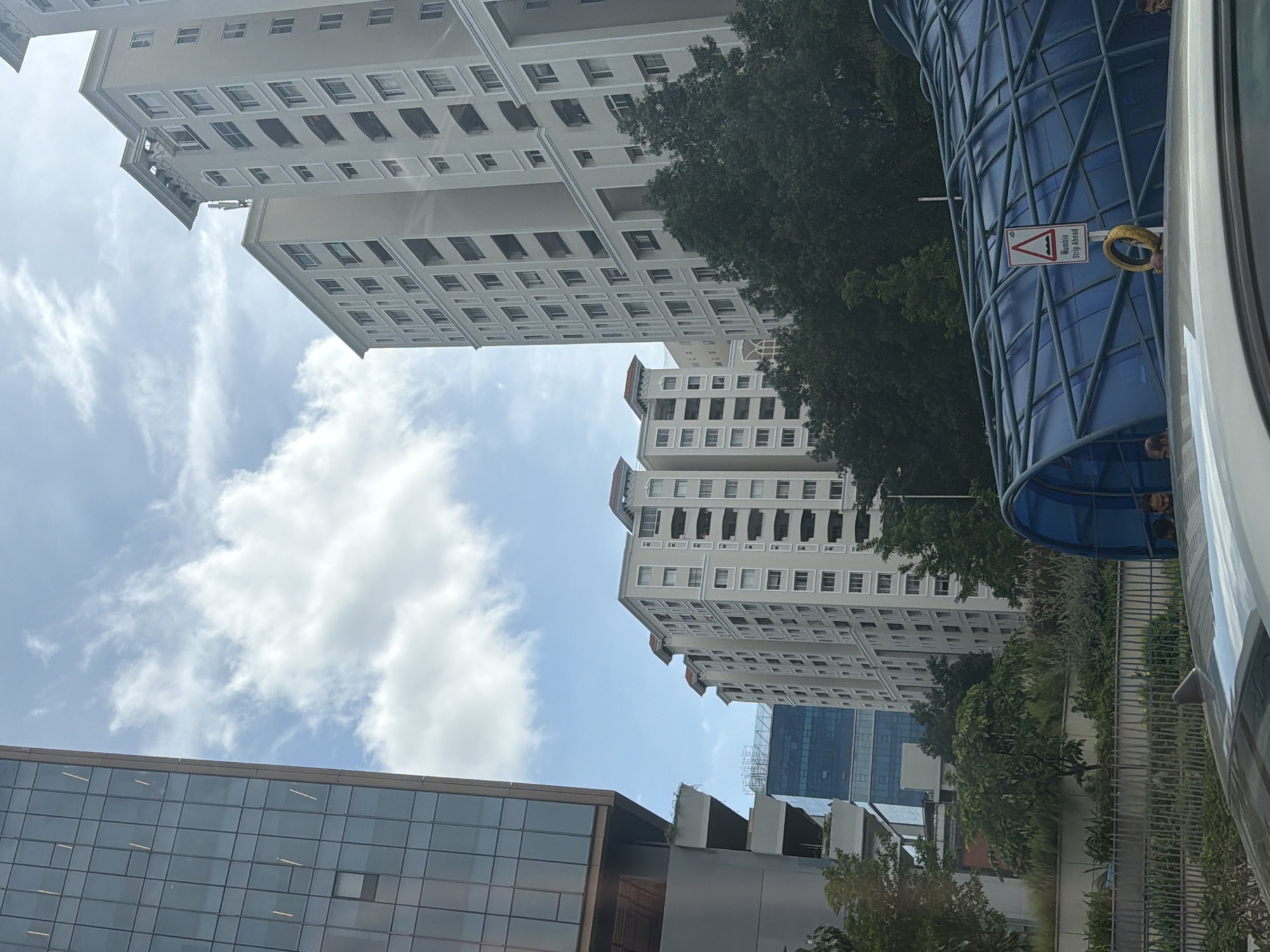 The image captures an urban scene in Hyderabad, India, featuring a cluster of modern buildings under a partly cloudy sky. On the left, a tall glass-fronted building with a copper-colored frame dominates the foreground. To its right, several multi-story residential buildings with light-colored facades and numerous windows are visible. The middle ground is occupied by lush green trees and foliage. In the foreground, a portion of a car's roof is visible, and to its right, a blue, semi-transparent tunnel-like structure is present. A road sign with the text "Hump/Strip Ahead" is affixed to a pole near the tunnel structure, along with a yellow, circular object. Two individuals, partially visible, are near the tunnel. The overall impression is of a well-developed urban environment with a mix of residential and commercial architecture, alongside green spaces.