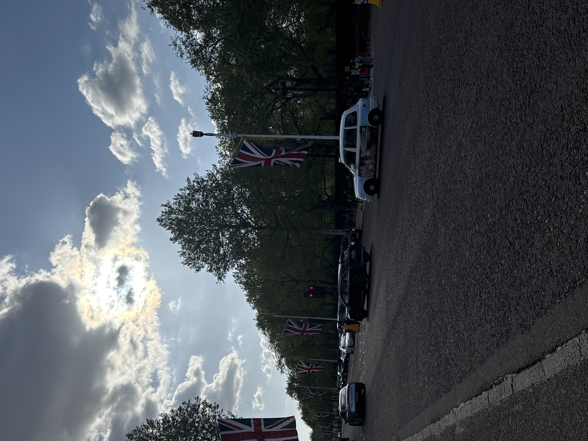 This image captures an outdoor street scene in London, United Kingdom, on a bright, partly cloudy day. The sun, partially obscured by cumulus clouds, is visible high in the sky, creating a radiant halo effect. A dark asphalt road, possibly a park road or main thoroughfare, stretches from the foreground into the distance.
Along the left side of the road, a dense line of mature green trees forms a natural canopy. Numerous Union Jack flags are prominently displayed, hanging vertically from poles positioned along the roadside, suggesting a celebratory period or national event.
A row of vehicles is either parked or stopped in traffic along the left side of the road. These include several traditional black London taxis and one distinctive light blue and white taxi, which features unreadable graphics or text on its side. Further down the street, a traffic light pole shows red lights, indicating stopped traffic. In the background, beyond the vehicles and trees, faint outlines of buildings or structures can be seen, along with a few indistinct figures on what appears to be a sidewalk or pedestrian area. The overall impression is one of a busy, yet scenic, urban setting with a strong sense of British national pride.