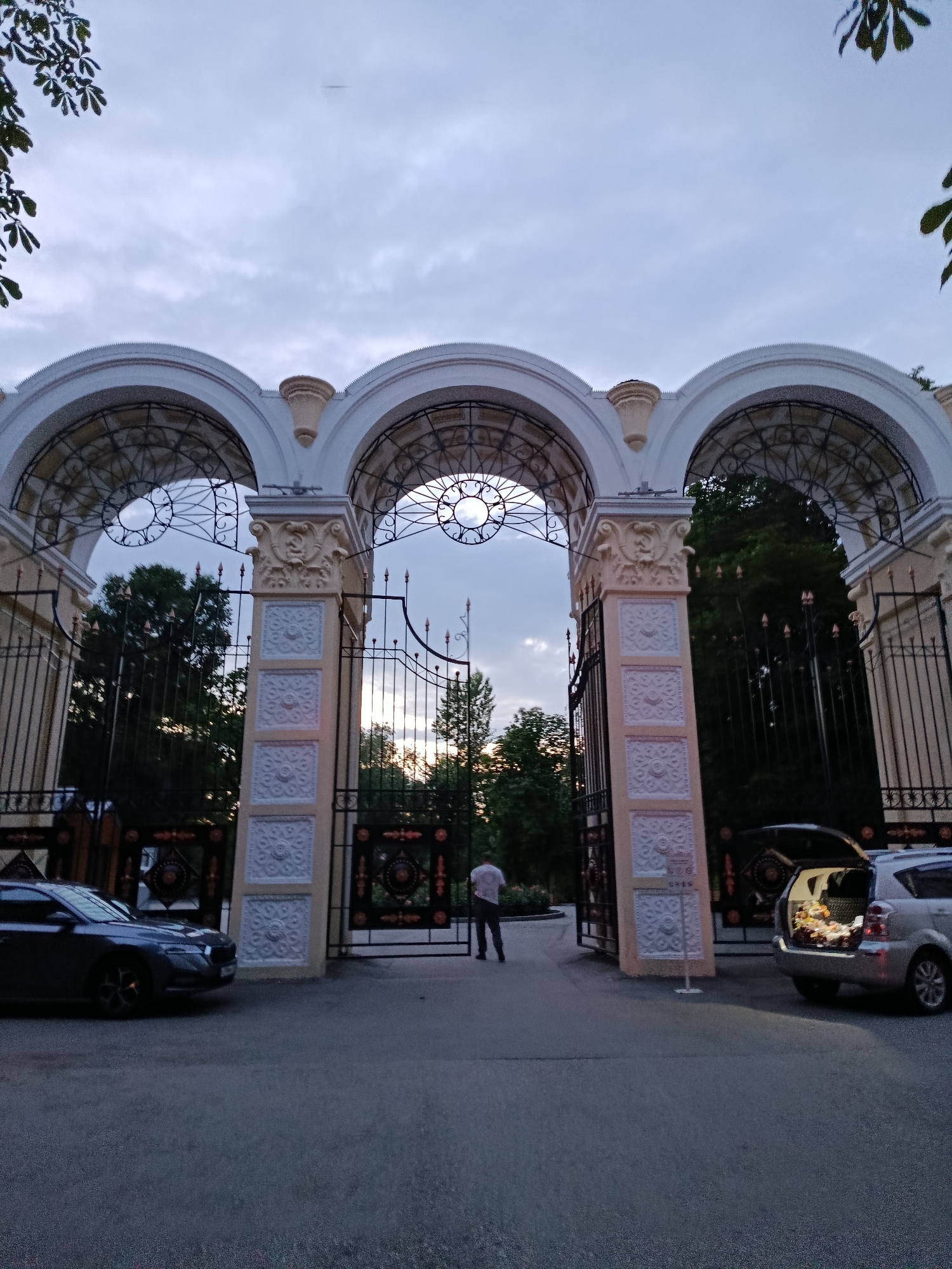 The image shows a wide view of a decorative arched entrance with three arches, reminiscent of a grand gateway. The entrance is adorned with intricate white plasterwork and metal grills. In front of the entrance, there is a paved road. To the left, a dark-colored sedan is parked. To the right, a silver minivan with its trunk open, revealing contents, is parked. A person in a white shirt and dark pants is standing in the middle of the road, facing away from the camera, near the entrance. The background features lush green trees and a cloudy sky, suggesting it might be dusk or an overcast day. The overall impression is one of an elegant and possibly historical entrance.
