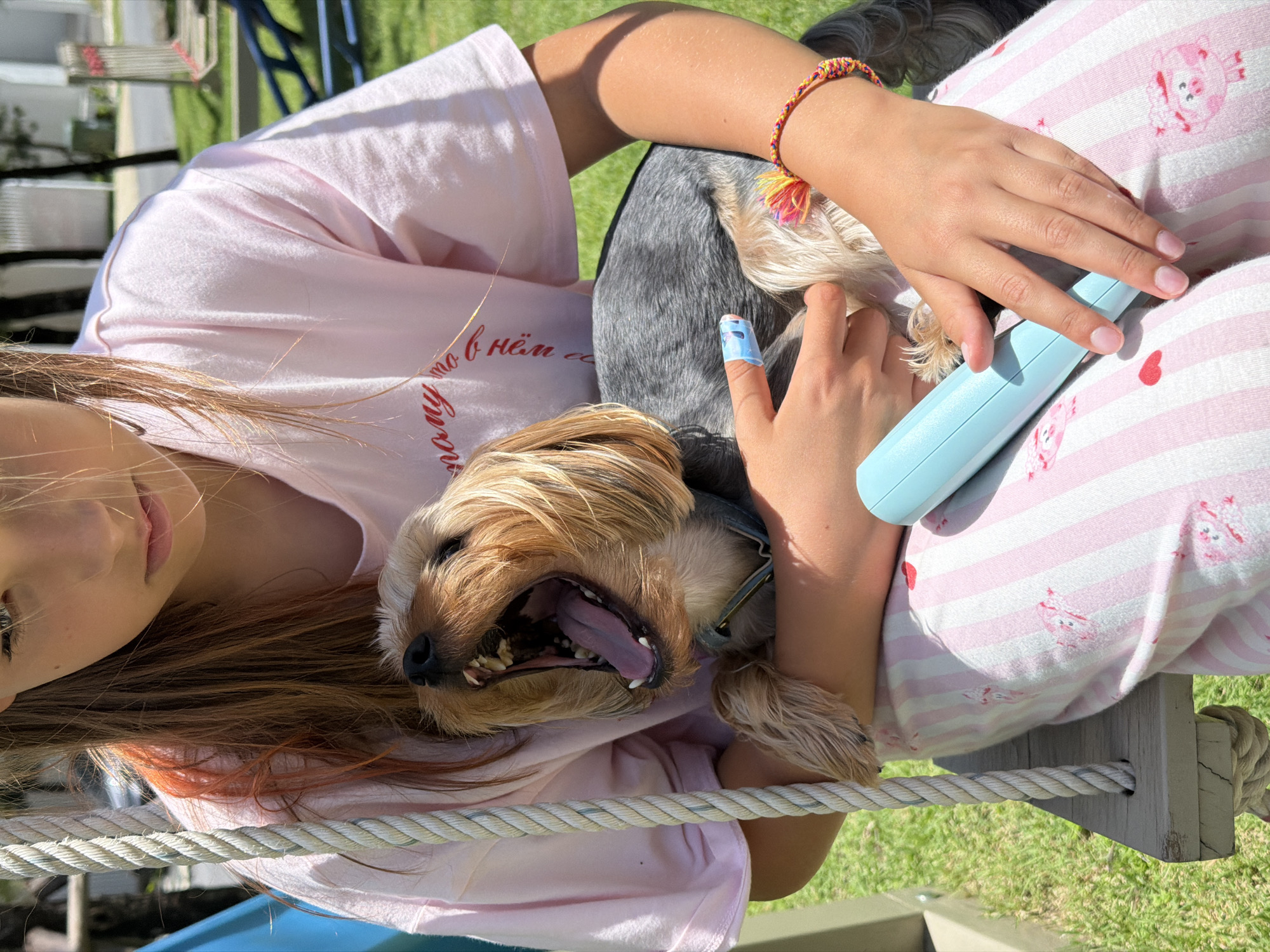 An outdoor close-up shot shows a young girl holding a small dog while sitting on a wooden swing. The girl, with blonde hair tinged with pink, is wearing a pale pink t-shirt and pink striped pajama bottoms. The dog, a Yorkshire Terrier, is yawning widely, revealing its teeth and tongue. A blue object, possibly a grooming tool, rests in the girl's lap. A colorful woven bracelet is visible on the girl's wrist. The background is blurred, showing green grass and trees. The location appears to be a residential backyard in Thep Krasatti, Thailand, indicated by the lush vegetation and sunlight typical of the region.