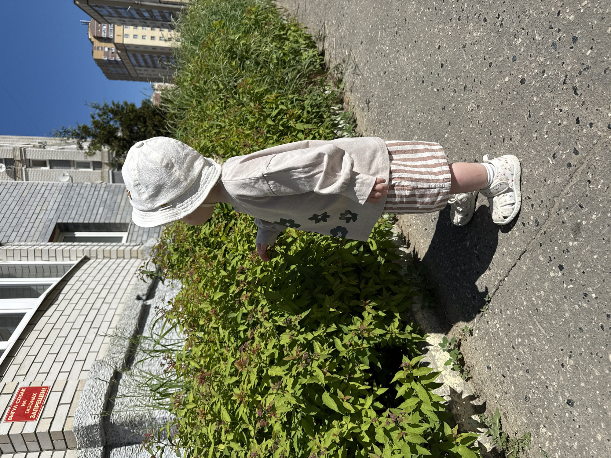 A child wearing a light beige hat, long-sleeved shirt with flower prints, striped shorts, and white sandals is standing on a sidewalk beside a lush green bush. The sidewalk is gray and textured. A building with a red sign is visible behind the bush. Taller buildings are visible in the background against a clear blue sky. The location is Vladimir, Russia.