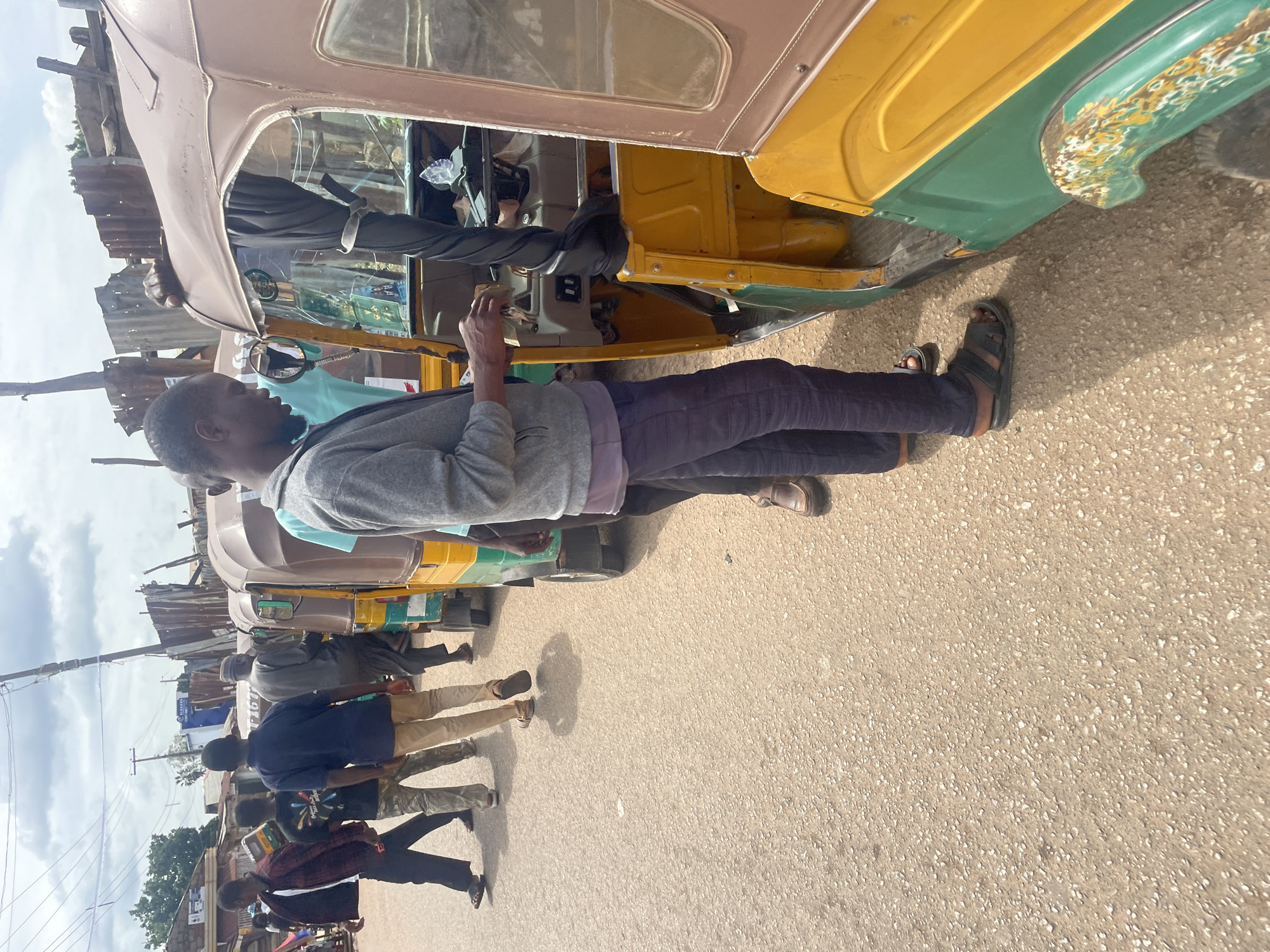 The image captures a street scene in Gura Top, Nigeria, with several people and auto-rickshaws (also known as Keke Napep). In the foreground, a man wearing a grey hooded sweater, dark pants, and sandals is standing next to a yellow and green auto-rickshaw, with his hands near the steering wheel. Behind him, other auto-rickshaws are lined up, some occupied by people. To the left, a group of men, some in casual clothing, are walking on the asphalt road. The background reveals a densely built area with corrugated metal structures and buildings, suggesting a busy urban environment. The sky is overcast with clouds.