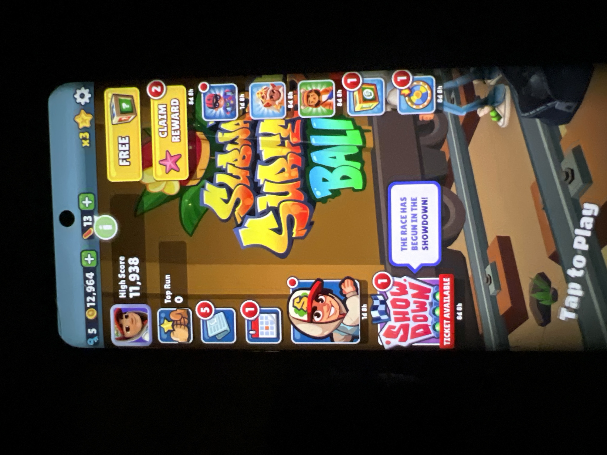 The image shows a screenshot of a mobile game, likely "Subway Surfers", displayed on a smartphone screen. The game interface features a character selection screen with various avatars and in-game items. The game appears to be themed around "Subway Surfers Bali". On the left side of the screen, there are icons for characters, high score, and other game features. On the right side, there are buttons for free rewards and claiming in-game items. A prominent banner at the bottom reads "Tap to Play". The background of the game shows a colorful, cartoonish environment with train tracks and a train. The smartphone is black, and the screen is illuminated, suggesting it is being actively used. There are no identifiable landmarks or elements that specifically place this image in Lagos, Nigeria.