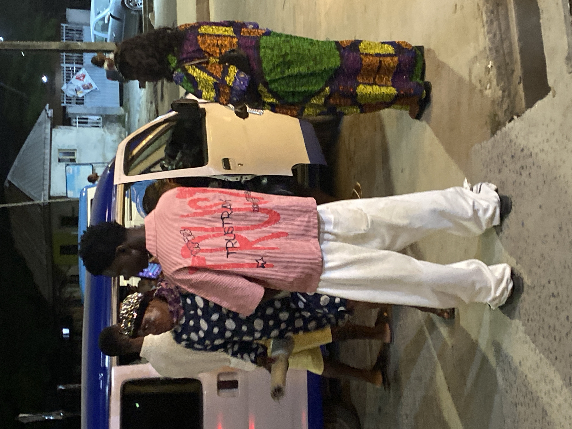 The image shows a group of people standing near a vehicle in Ado, Nigeria. One person is wearing a colorful, patterned outfit, while another wears a pink shirt with the word "TRUST" on it and white pants. Additional individuals are dressed in casual attire. The vehicle is partially visible and appears to be a van-type vehicle with blue accents. The background shows buildings and street lights, suggesting an urban setting. The scene appears to be at night or in low light conditions.