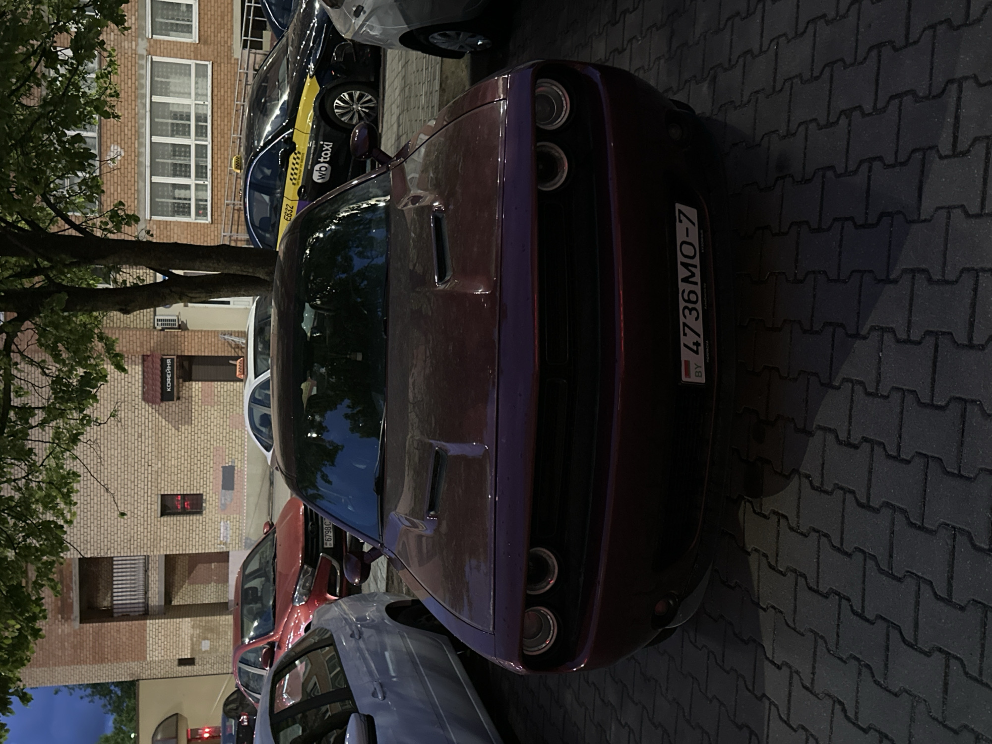 The image depicts a plum-colored Dodge Challenger parked on a paved street in Minsk, Belarus. The car's license plate reads "4736 MO-7" and includes the country code "BY". Behind the Challenger, there are other parked vehicles, including a taxi with the markings "wb taxi" and the license plate "E832", and a red car with the license plate "6798 C." A building can be seen in the background, with the word "КОФЕЙНЯ" (Coffee Shop) visible on one of the signs. There is a tree to the left of the building. The pavement is made of interlocking concrete blocks. The image was taken at dusk.