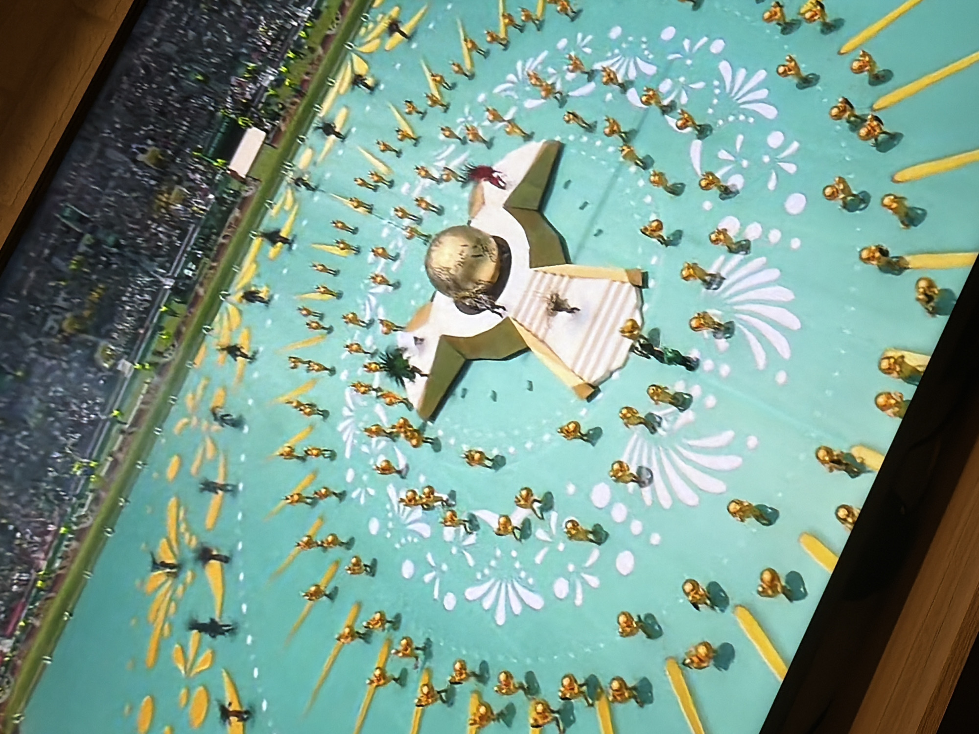 The image depicts an aerial view of a large outdoor event, possibly a ceremony or performance, taking place in a stadium. The ground is a bright teal color, decorated with white floral patterns and scattered yellow shapes that resemble petals or rays. Numerous small figures, appearing to be performers dressed in gold costumes, are positioned across the teal surface, many of them in organized rows or formations.
In the center of the field, a large, stylized, star-shaped structure is visible, topped with a golden sphere. Performers are also present on and around this central structure. In the background, the stadium seating area is filled with a large crowd of spectators, some of whom are holding green and white flags.
The scene strongly suggests a cultural event or a sporting ceremony, such as an opening or closing ceremony. Given the context, it's possible this is a shot from a broadcast or recording of such an event. However, the provided image does not contain any visual cues that would allow for the identification of a specific city or location, such as Pazardzhik, Bulgaria, nor does it offer any clues about the nature of the event beyond its visual spectacle.