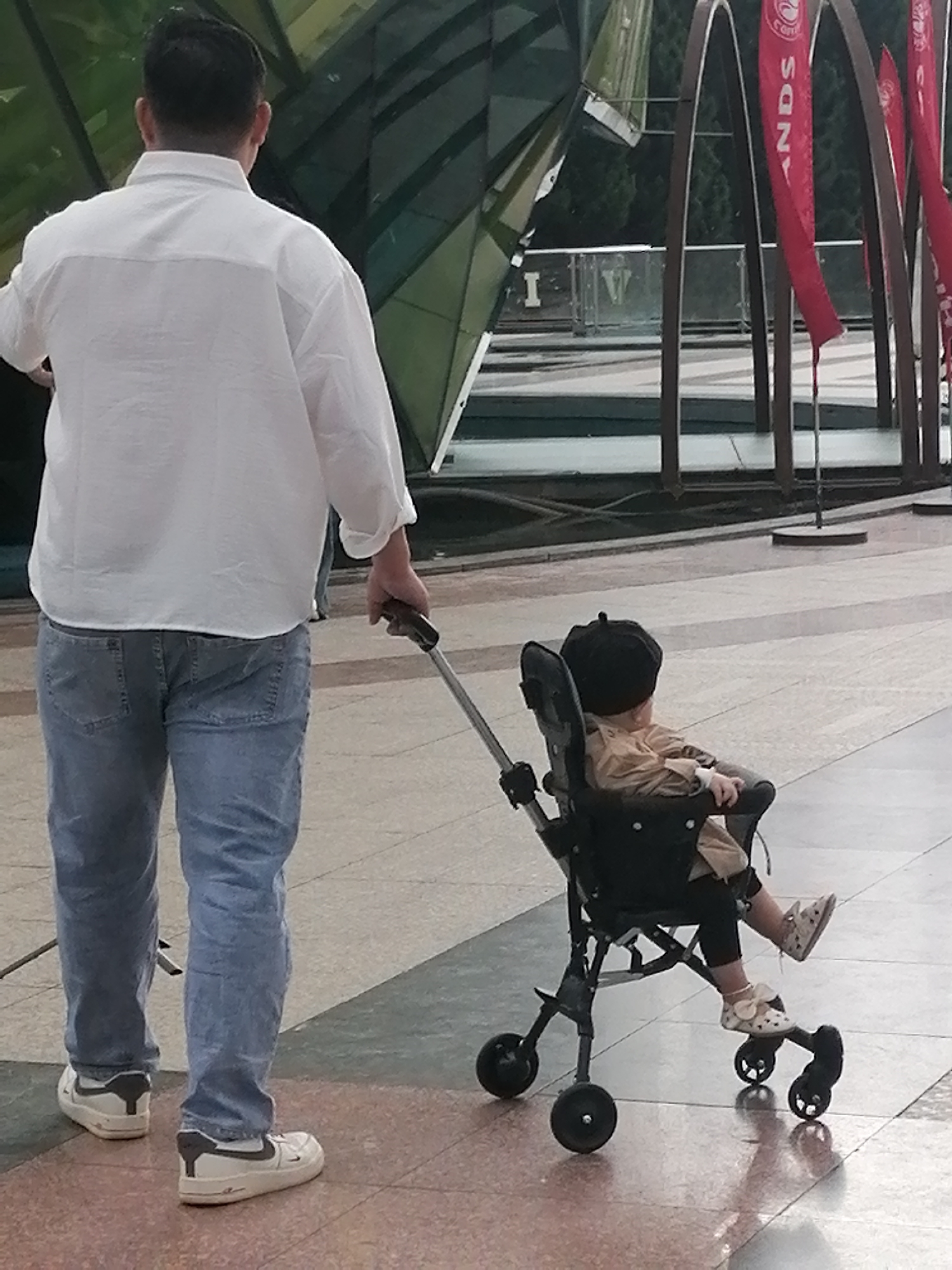 A man is pushing a stroller with a child in it. The man is wearing a white shirt and blue jeans. The child is wearing a tan jacket and a black hat. The stroller is black. The background appears to be a plaza or park area with some modern architecture. The image was taken in Da Lat, Vietnam.