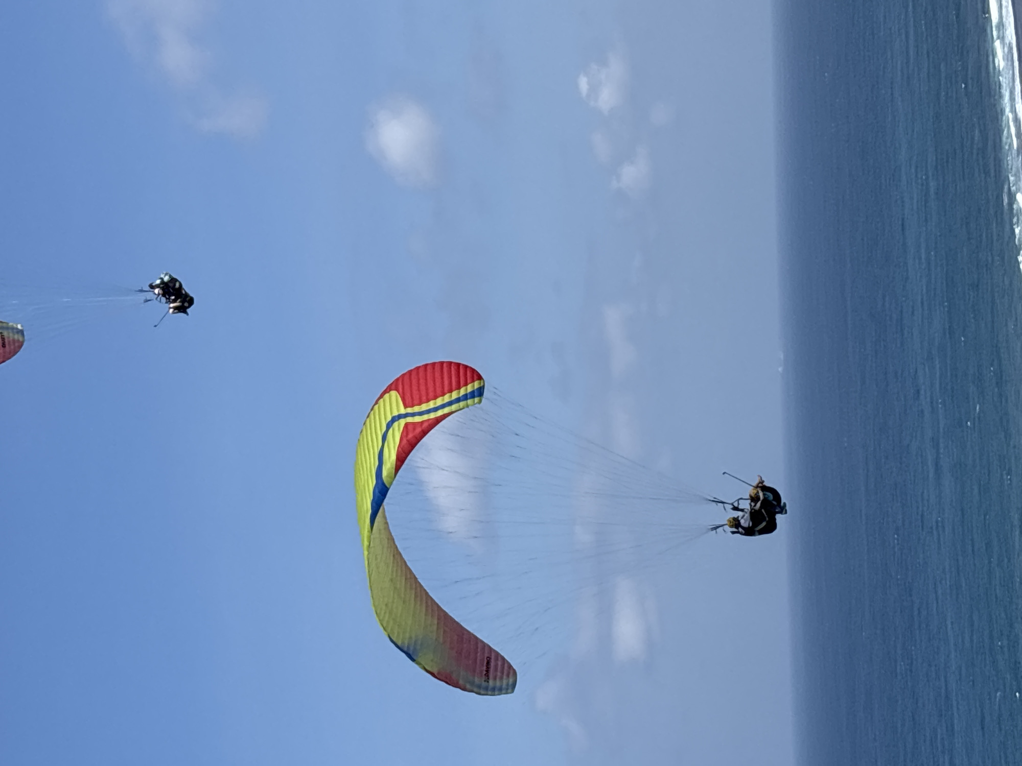 This media file captures a paragliding activity over a body of water, likely the ocean, in Kuta Selatan, Indonesia. The scene depicts at least three paragliders suspended in the air against a bright blue sky dotted with a few wispy white clouds.
The primary focus is on a large, colorful paraglider, predominantly yellow with red and blue stripes, soaring prominently in the foreground. Attached to this paraglider are two individuals, appearing to be in tandem. Their figures are small against the vastness of the sky and the paraglider's wing. Further in the distance, two other paragliders are visible, also appearing to be in tandem, smaller and more to the left of the frame.
The activity appears to be recreational or sport-oriented, with the paragliders enjoying the flight. The weather seems to be fair and sunny, indicated by the clear blue sky and the bright lighting. The presence of the expansive ocean to the right suggests a coastal location, consistent with Kuta Selatan being a popular destination for such activities. The time of day appears to be daytime, likely late morning or early afternoon, given the intensity of the sunlight.
One paraglider wing shows some visible text, though it is difficult to decipher completely due to the resolution and angle. However, parts of what looks like a brand name or model number might be present. No explicit emotions are conveyed, but the activity itself suggests a sense of adventure and freedom. The overall impression is one of a serene yet exhilarating experience of soaring through the sky above the ocean.