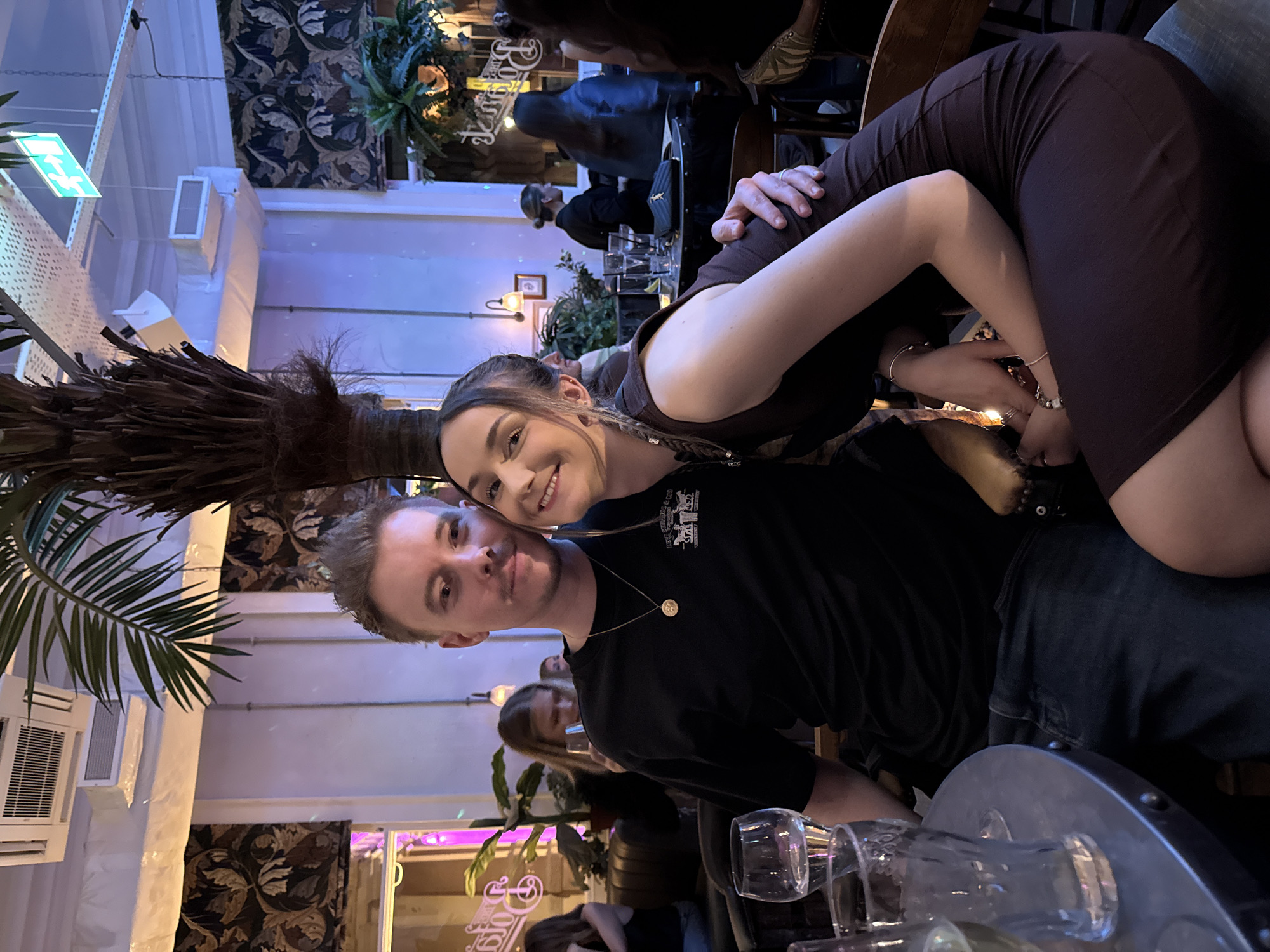 A vertical, full-frame photograph shows a couple posing for a selfie in what appears to be a bar or restaurant. The man, with short blond hair and wearing a black t-shirt and jeans, is in the foreground, partially obscured by glassware on the table. He has a gold pendant necklace. The woman, with braided hair, leans in close with her arm around him, both smiling at the camera. She wears a brown dress.
In the background, other patrons are visible seated at tables. The environment is dimly lit with a mix of natural and artificial light, contributing to a relaxed atmosphere. Decorative plants and patterned wallpaper add to the establishment's ambiance. There appears to be an air conditioning unit. The establishment is identified as being in the city of Ipswich, United Kingdom.