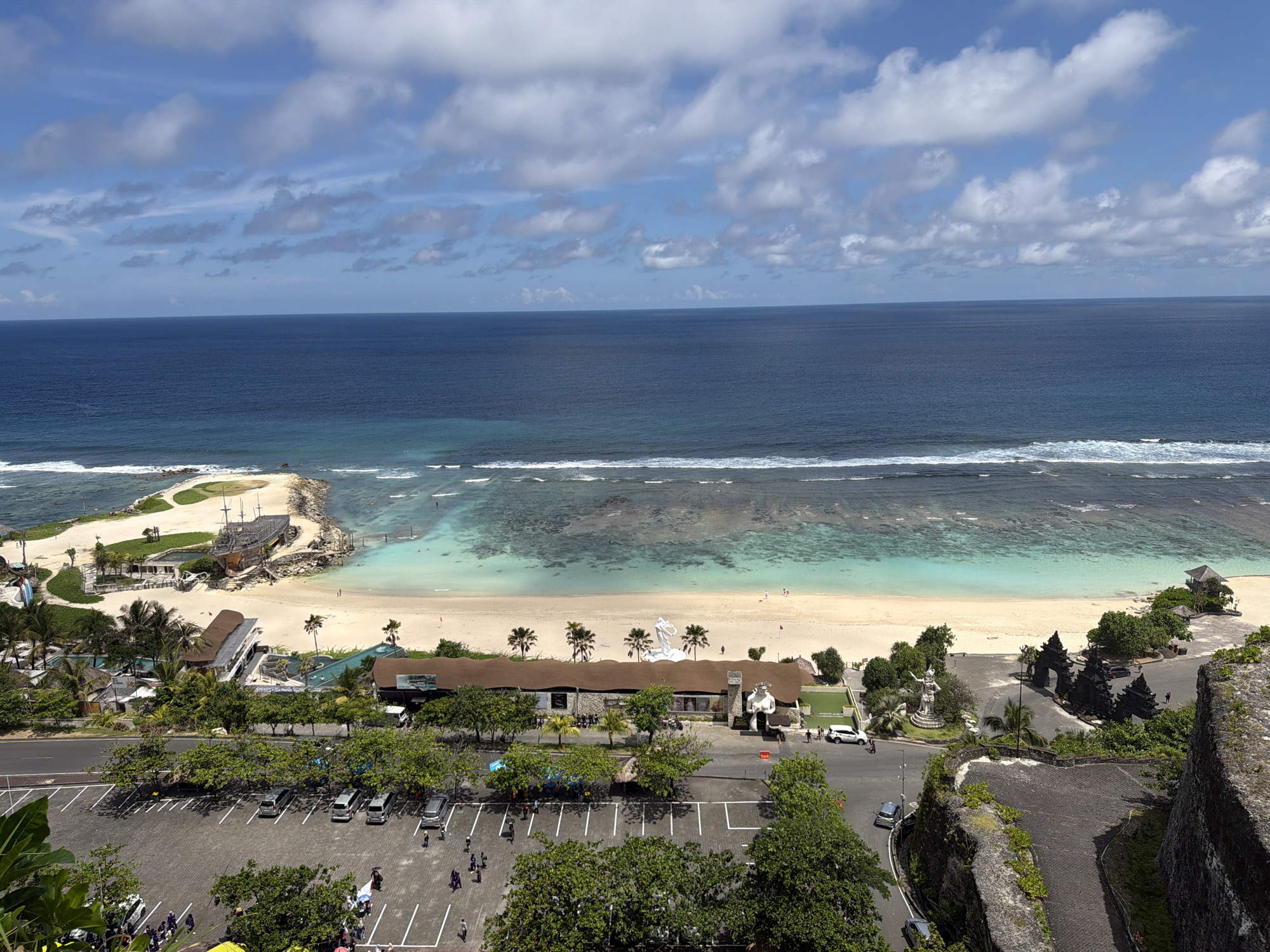 The image shows a high-angle view of Melasti Beach, located in Ungasan, Indonesia. The scene includes the ocean, beach, and surrounding infrastructure. The ocean extends to the horizon with a band of waves breaking along the shore. The beach is sandy with some rocky areas visible in the water.
A beached pirate ship is positioned prominently on the sand near the left side of the beach. Behind the beach, there are buildings and palm trees, along with an adjacent parking lot filled with vehicles and people. Some people are walking around in the parking lot. A road is visible below the parking lot and to the right of the parking lot. The sky is blue with scattered clouds.