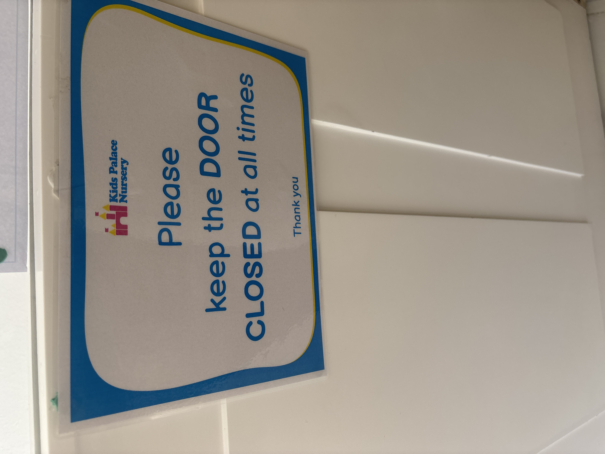 A sign with blue and yellow trim and a cartoon castle logo that reads "Kids Palace Nursery" is attached to a white wall. The sign contains the text "Please keep the DOOR CLOSED at all times" and "Thank you." The image is a close-up shot of the sign, with the lower half of the sign and the white wall dominating the frame. The sign appears to be laminated. The environment is indoors, likely in a nursery or school setting, given the sign's content. There are no people or actions depicted in the image. The location is not specified in the image or its context.