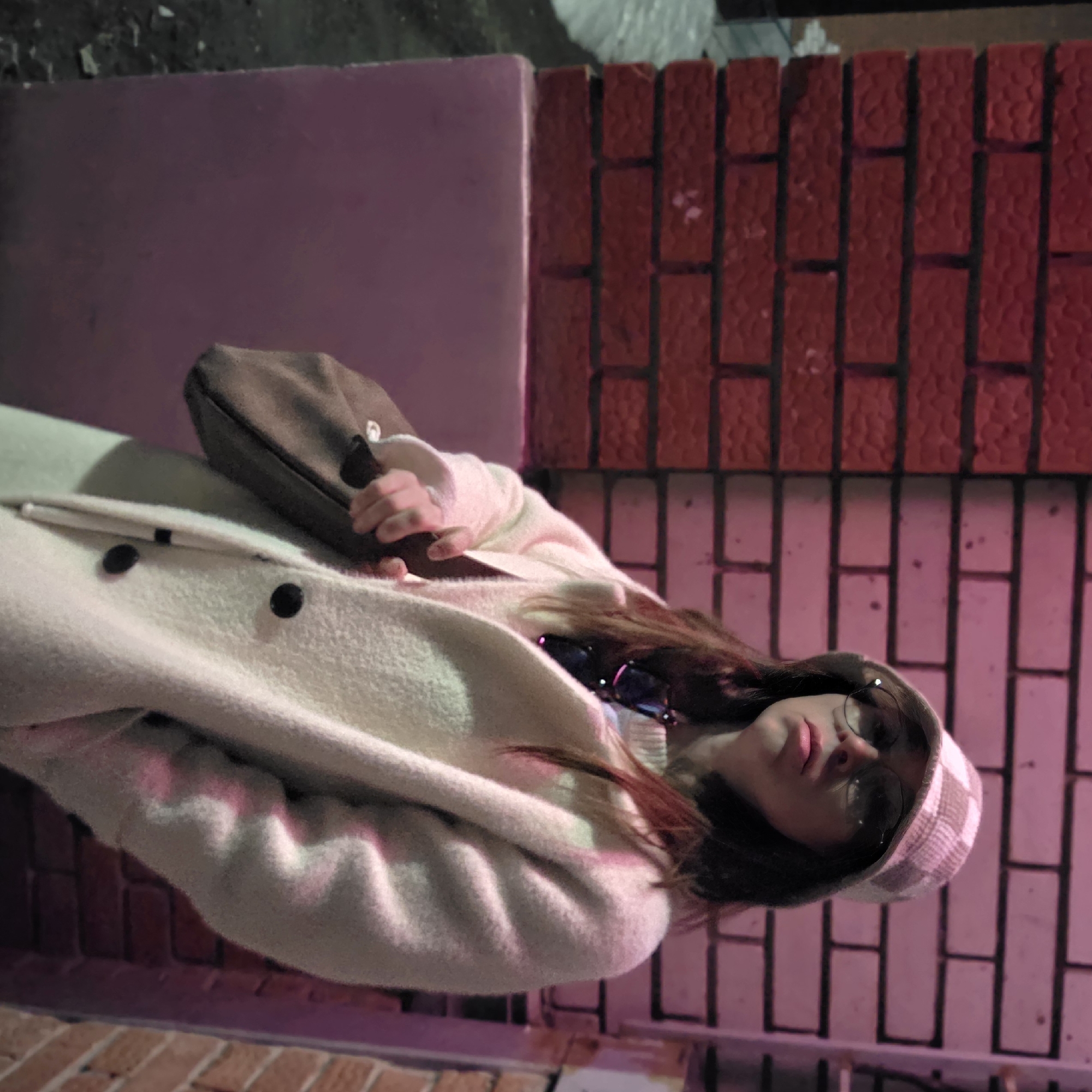 An adult female is depicted standing against a brick wall in Chaikovsky, Russia, under artificial lighting, likely at night. She is positioned slightly to the right of the frame, looking slightly upward and to her left (viewer's right). She wears a light-colored, plaid-patterned bucket hat, round-framed eyeglasses, and a long, cream-colored, fuzzy textured coat with visible dark buttons. Dark hair is visible extending from beneath her hat. Dark-framed sunglasses are hanging from her neck, partially visible beneath her coat. She holds a brown shoulder bag on her left side, gripping the strap with her left hand. Her right hand is placed into her coat pocket. The background features a brick wall, horizontally divided in color. The section to the viewer's left is dark red brick. The section behind the subject and to the viewer's right is also brick, illuminated with a distinct pinkish-purple hue. A smooth, pinkish-purple painted lower wall or base is visible beneath the red brick on the left. A dark, textured surface, possibly pavement or snow, is visible on the far left.