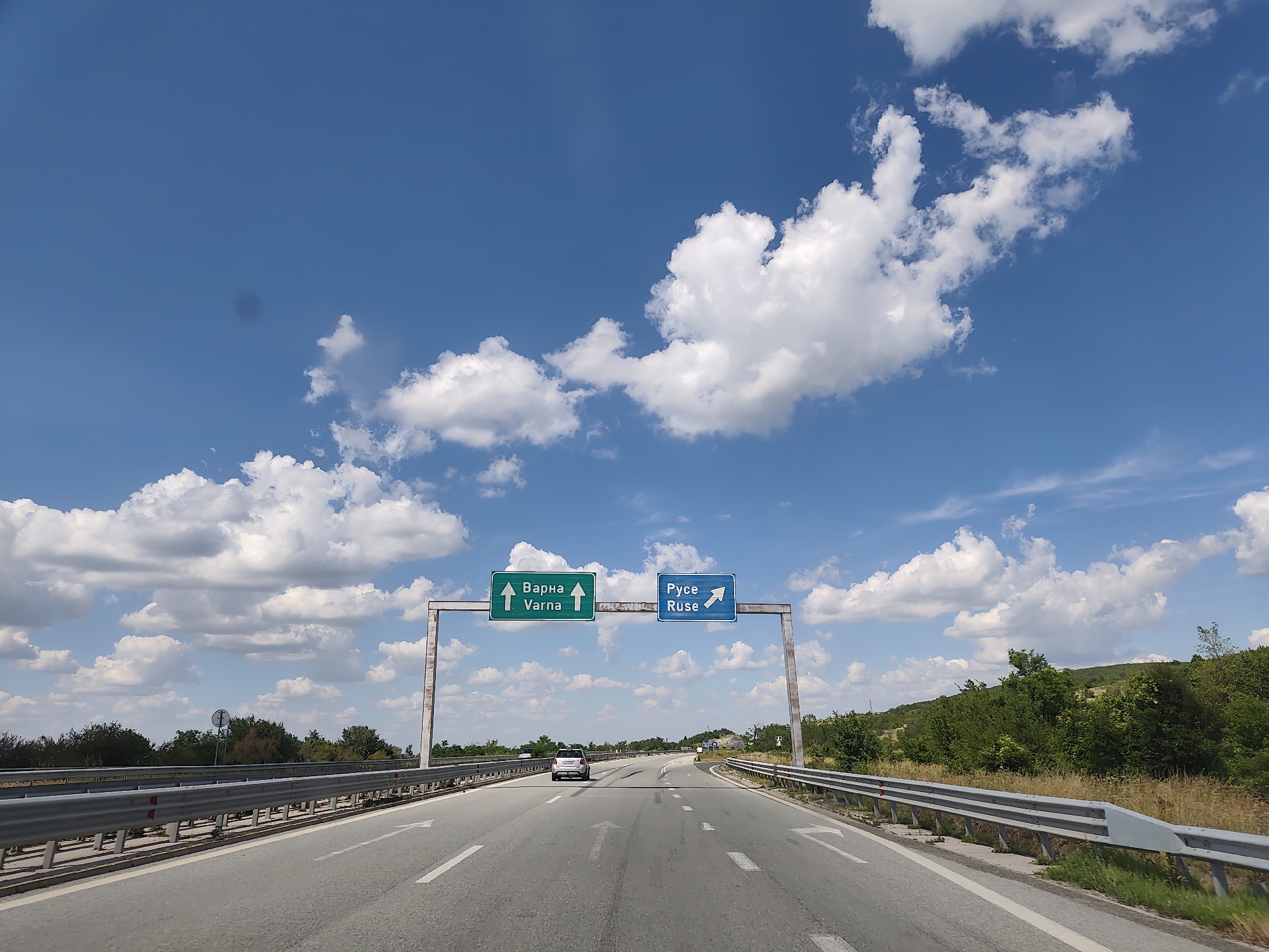 This image depicts a highway scene under a bright blue sky with scattered white clouds. A split-view overhead highway sign is centrally located. The left sign is green with white text and an upward arrow, indicating "Varna" in both Cyrillic and Latin script. The right sign is blue with white text and an upward-right arrow, indicating "Ruse" in both Cyrillic and Latin script. The highway has multiple lanes marked with white arrows. A silver car is visible in the left lane ahead. A metal guardrail runs along the right side of the road, and to the right of the guardrail, there is dry grass and vegetation, followed by a tree-covered hillside. To the left of the road, there is also a guardrail and a line of trees and bushes. The overall environment suggests a rural or semi-rural highway setting. This location is identified as being on the highway near Yablanitsa, Bulgaria, likely referring to the A2 Hemus highway which connects Sofia to Varna and passes near Yablanitsa, and also serves the route towards Ruse.