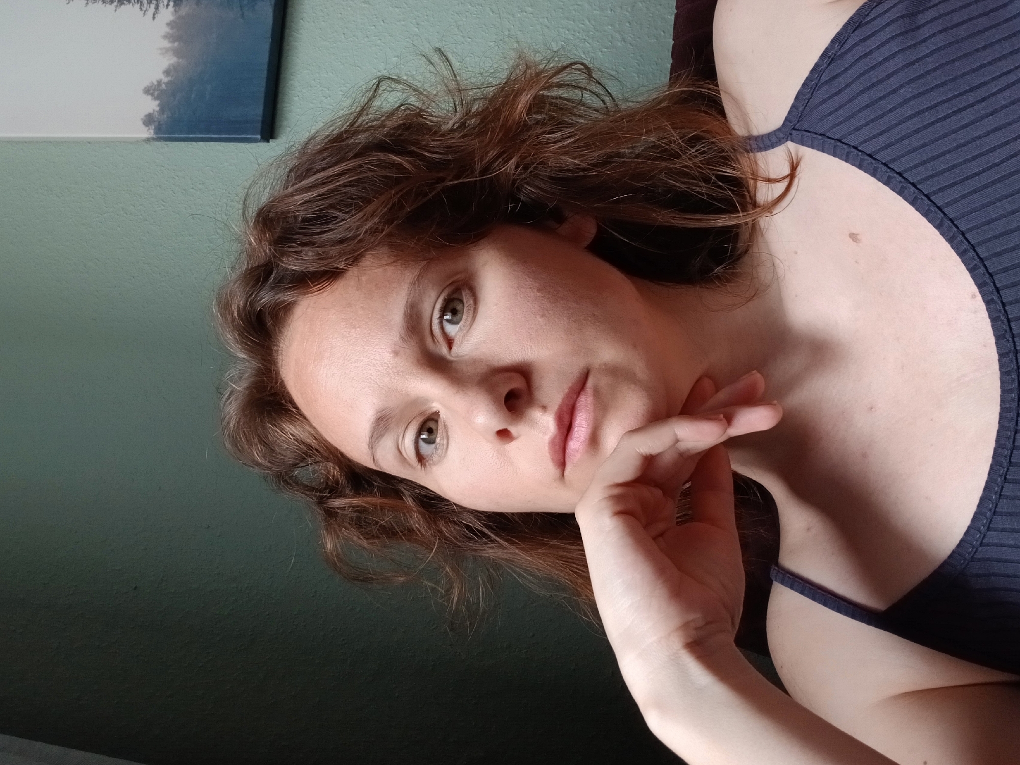 A close-up, front-facing portrait of a young Caucasian woman with light skin, brown hair, and green eyes is shown indoors, with her left hand resting on her chin, looking directly at the camera. She is wearing a dark blue ribbed tank top. The background consists of a textured, light teal wall, and to the right, a portion of a framed picture depicting a misty forest scene is visible. The lighting suggests an indoor setting, possibly a room. The image does not contain any elements that indicate its location in Yekaterinburg, Russia.