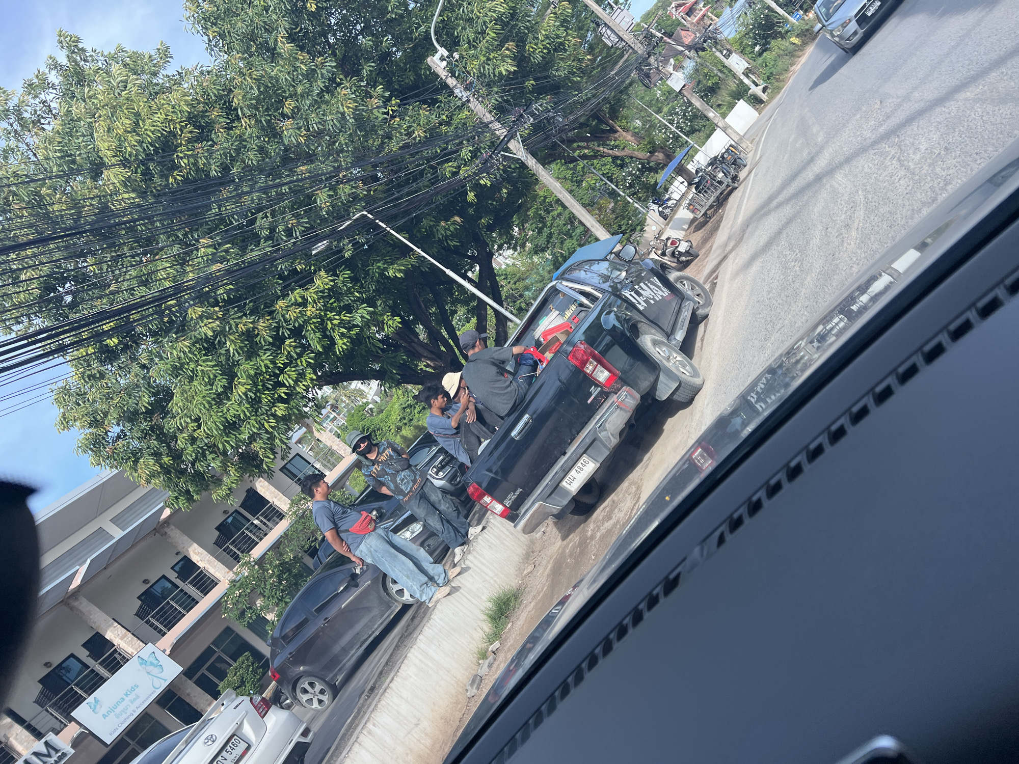 The image, taken from inside a vehicle, shows a street scene in Baan Plai Laem, Thailand. A group of people stand near a black Isuzu D-Max pickup truck parked on the side of the road. The truck's license plate reads "บพ 4846." To the left of the truck, a dark sedan is parked, partially obscuring a white car with license plate "วง 5460." A building with a sign that reads "Anjuna Kids Kids Clothing & Accessoires" can be seen in the background. Above, power lines intersect the sky. Vegetation and street scenes are visible in the background.