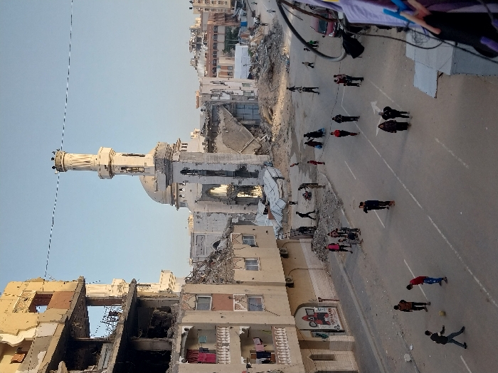 The image captures an aerial perspective of a heavily damaged urban street scene during daytime. Many multi-story buildings lining the street show extensive structural damage, with walls collapsed, exposing interiors, and large piles of rubble and debris accumulated on the ground and along the roadside.
In the mid-ground, a prominent mosque stands, featuring a tall, slender minaret with a crown-like top and a white dome. The mosque also exhibits visible damage to its structure, though the minaret appears largely intact. The sky is clear and bright blue, indicating fair weather.
A paved road runs through the center of the scene, with faint white lane markings and a white directional arrow visible on its surface. Numerous individuals, appearing to be adults and children, are scattered across the street. They are seen walking, standing in small groups, or individually, navigating the environment. In the lower right foreground, a group of young people or children appears to be engaged in play, with one individual seen with a ball.
Overhead wires or cables crisscross the upper portion of the frame. On the side of a partially damaged building in the lower-left, a mural is visible. It depicts a figure resembling a guard from the TV series "Squid Game" (wearing a red suit and mask). Below the figure, some Arabic text is written, which appears to spell out "سكويد جيم" (Squid Game).
The scene portrays daily life continuing amidst a landscape of significant destruction, suggesting an urban area that has experienced considerable upheaval or conflict. The elevated viewpoint provides a broad overview of the extent of the damage and the presence of people in their immediate environment.