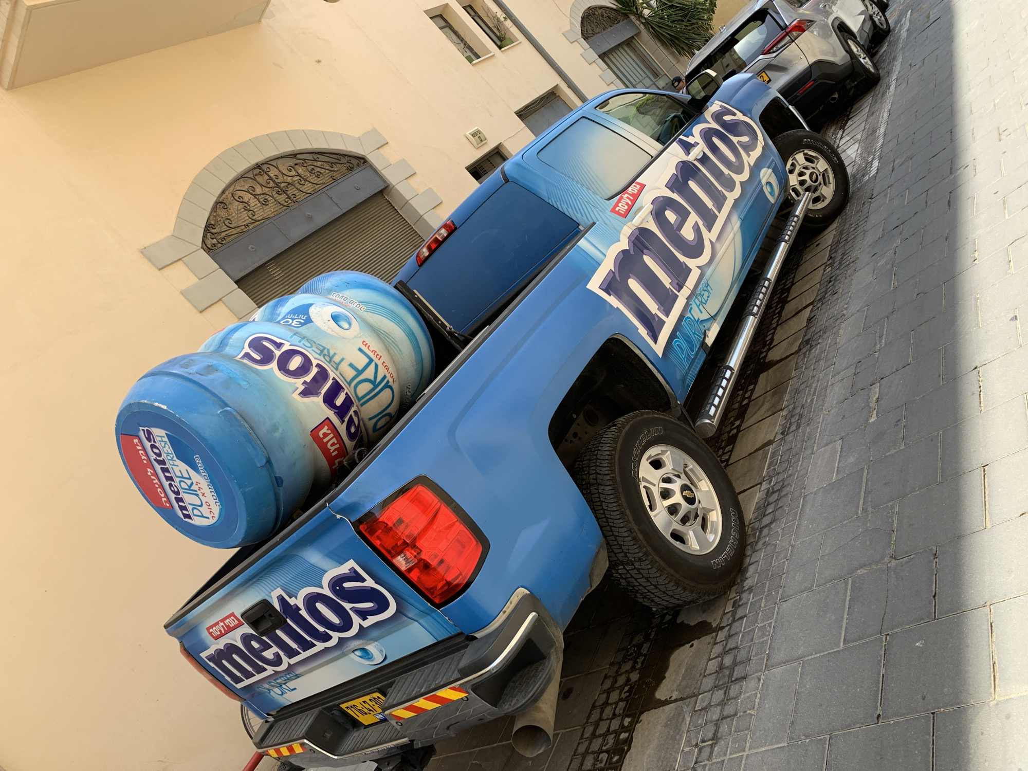 A blue pickup truck is parked on a brick-paved street in Tel-Aviv, Israel. The truck is emblazoned with "Mentos Pure Fresh" advertising, including oversized Mentos gum containers in the truck bed. The truck bed and side panels also feature Mentos logos. A silver car is parked ahead of the truck. The surrounding environment includes buildings with arched windows and window gratings.