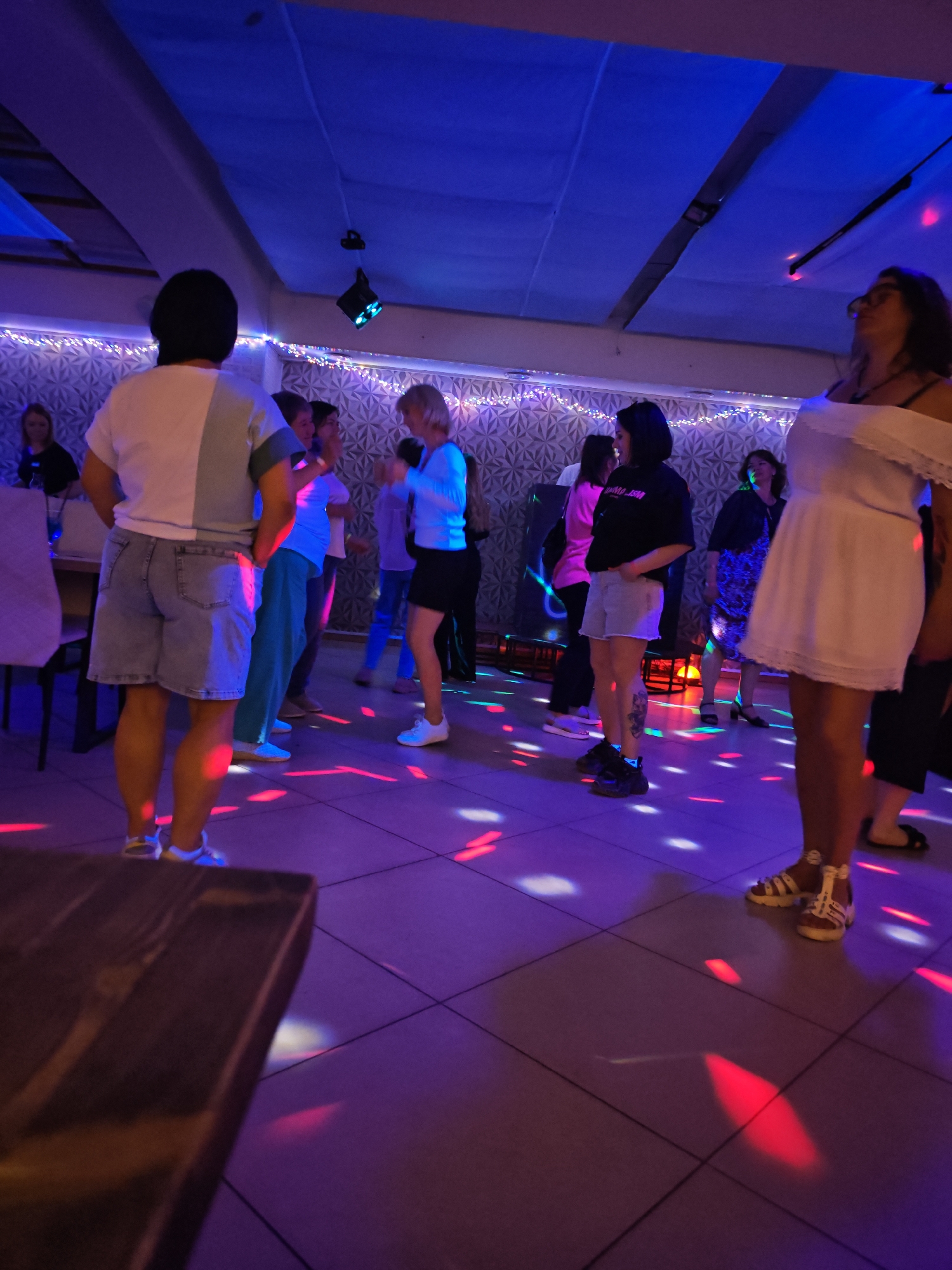 People are dancing in a room with tiled floors illuminated by red and blue disco lights. Most of the people are facing forwards and are wearing casual clothes. The room is decorated with lights along the top of the walls. The location is in the city of Chaikovsky, Russia.