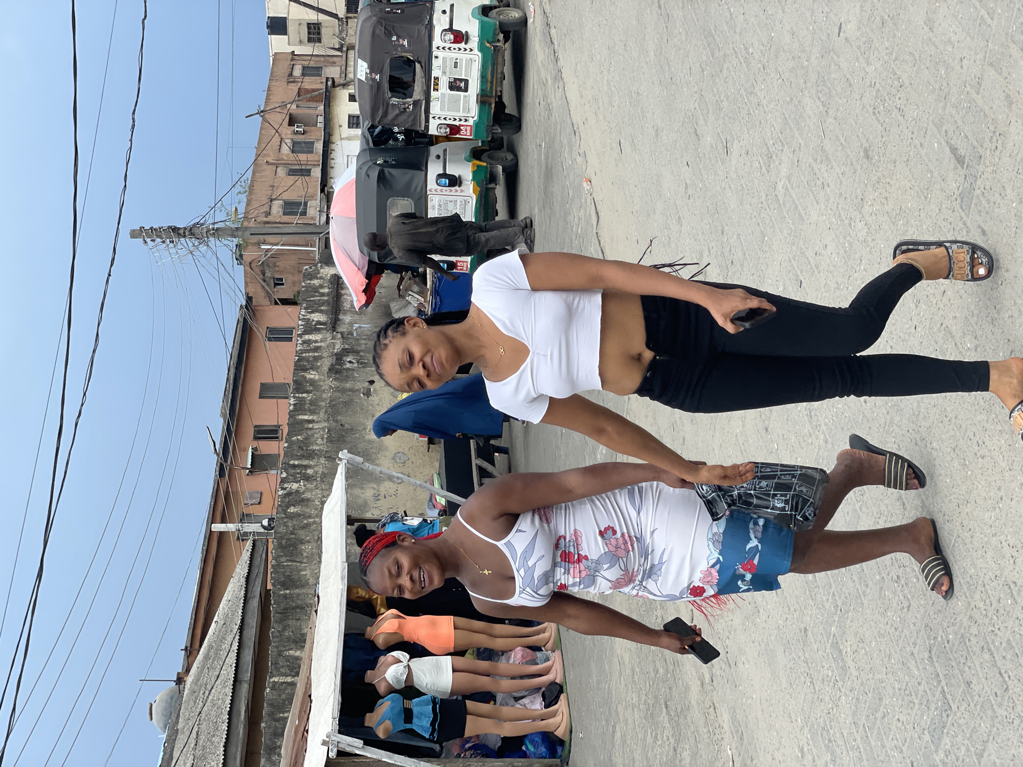 Two women are standing on a concrete surface, possibly a street. The woman in the foreground wears a white dress with floral patterns and a red headscarf; she is holding a cell phone in her right hand. Beside her are several mannequin legs wearing different colored outfits. The woman in the background is wearing a white crop top and black pants, and also holding a cell phone in her right hand. Behind them, there are several auto-rickshaws parked on the side of the street and a man standing nearby. In the background, there is a building and a utility pole with many wires. The location appears to be a city street.