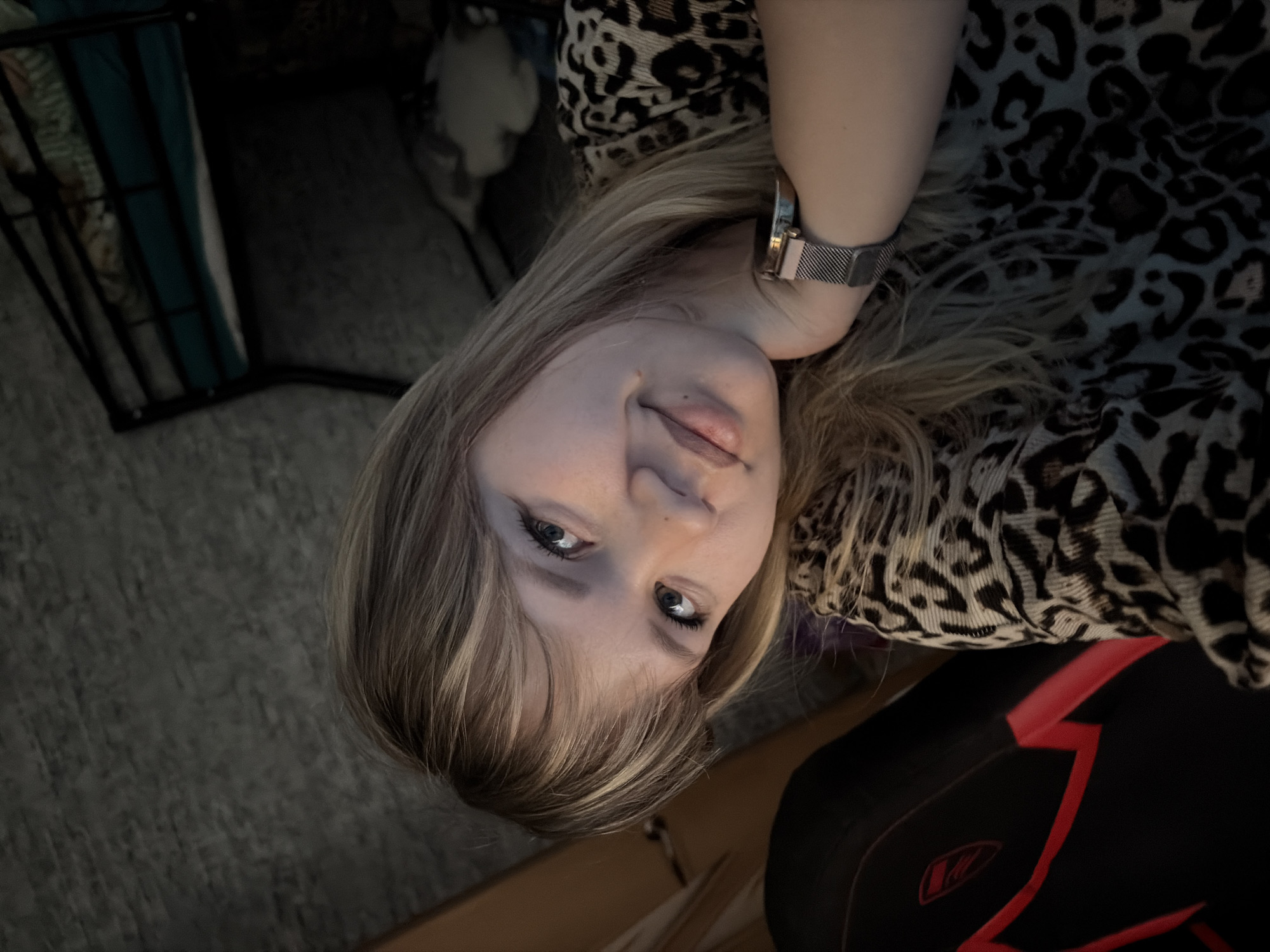 A Caucasian woman is seen from a low angle, lying on her back. She has fair skin, blonde hair with dark roots, and blue eyes. She is wearing a leopard-print top and a watch on her left wrist.
She appears to be indoors. The background includes a carpet, a black and red chair, and a black frame structure with items inside.
There is no information in the image that indicates it was taken in Chaikovsky, Russia.
