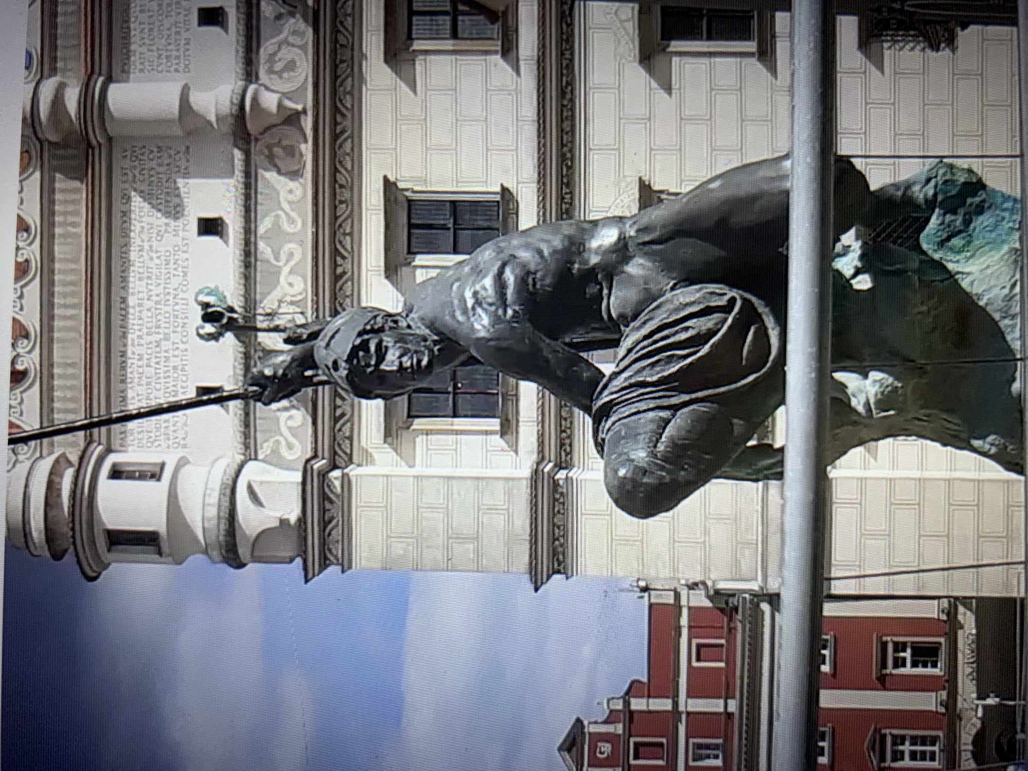 The image shows a close-up of a statue in a city square. The statue appears to be made of dark metal, possibly bronze, and depicts a muscular man in a crouching or kneeling position. He is wearing a helmet and a draped garment. The statue is mounted on a rocky base, which has a greenish patina. In the background, there is a building with white facade and patterned brickwork, featuring several windows. To the left, a red building with white trim and multiple windows is visible. The sky above is blue with some clouds. The statue is positioned in front of a metal railing. There is no visible indication of the location being Lymanka, Ukraine, nor any indication of people interacting with the statue or engaging in any actions.
