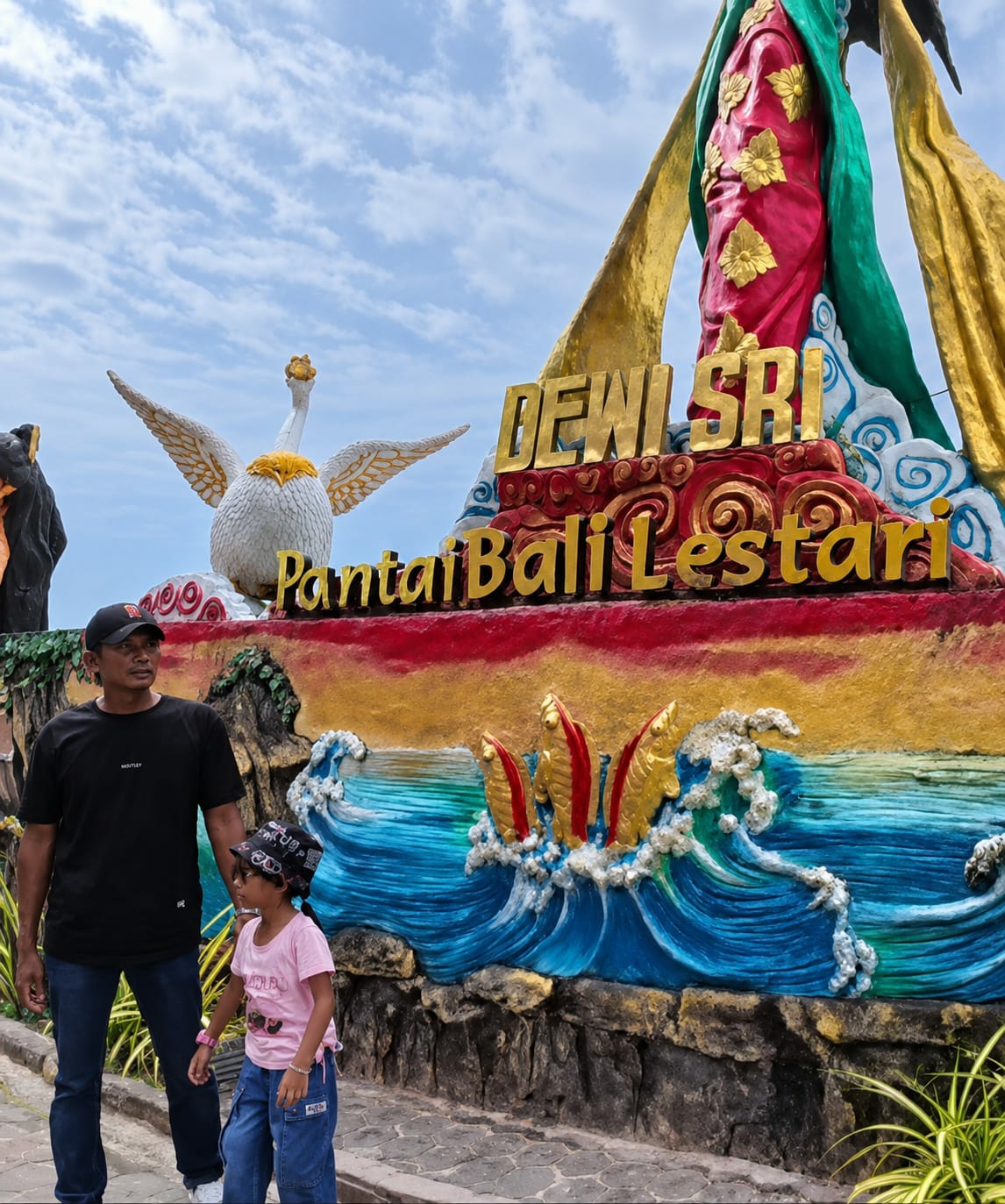 The image depicts a father and daughter standing in front of a colorful mural and statue, identified as being located at Pantai Bali Lestari in Kota Pari, Indonesia. The mural features a large, ornate statue of a figure, possibly Dewi Sri, adorned in red and green robes with gold accents. Below the statue, the words "DEWI SRI" are emblazoned in gold lettering, followed by "PantaiBali Lestari" in smaller gold letters. The mural itself depicts vibrant blue waves crashing against a rocky shore, with stylized golden fish or sea creatures emerging from the water. To the left of the main statue, a white swan-like sculpture with wings spread wide is visible. The father, wearing a black t-shirt, black cap, and blue jeans, stands to the left of the child. The daughter, dressed in a pink t-shirt and blue jeans, wears a patterned bandana on her head. They are both looking towards the mural and statue. The background shows a cloudy blue sky. The ground beneath them is paved with cobblestones.