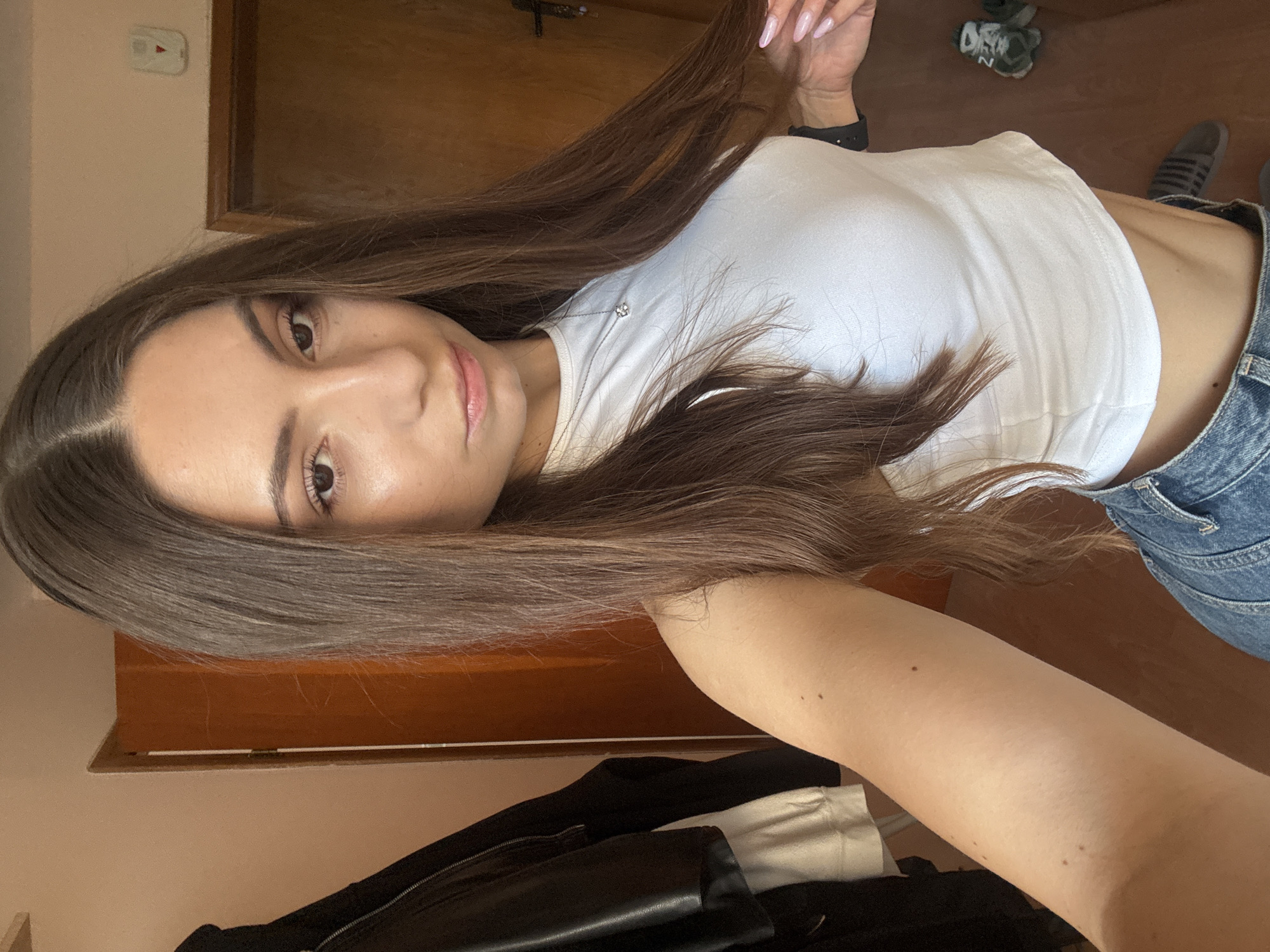 A young woman with long, brown hair is taking a selfie indoors. She is wearing a white crop top and blue jeans. Her skin is light-toned with several small moles visible on her arm. She has a silver necklace with a small pendant. Her fingernails are painted pink. In the background, there is a wooden door, a black jacket hanging on a rack, and a pair of sneakers on the floor. The lighting suggests it is daytime, possibly near a window. The scene is domestic and appears to be within a residential building. There is no discernible information to pinpoint the exact location within Varna, Bulgaria.